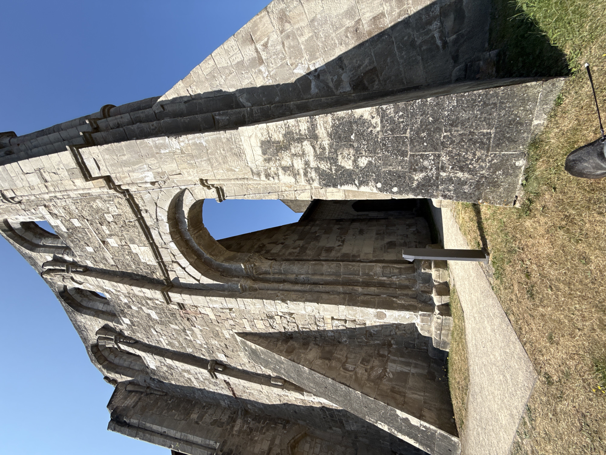 The image shows a low-angle shot of the ruins of Walkenried Abbey in Walkenried, Germany. The stone structure, built from large, rough-hewn blocks, is weathered and shows signs of age. The sky is a clear, bright blue. In the foreground, a path of gravel leads towards the ruins, bordered by dry, brown grass and some patches of green. To the right, a dark, angled shadow is cast across the stone wall. A black boot with a thin white-tipped object extending from it is visible in the bottom right corner of the frame, positioned on the grass. The ruins feature an archway and several empty arched window openings along the top of the wall.
