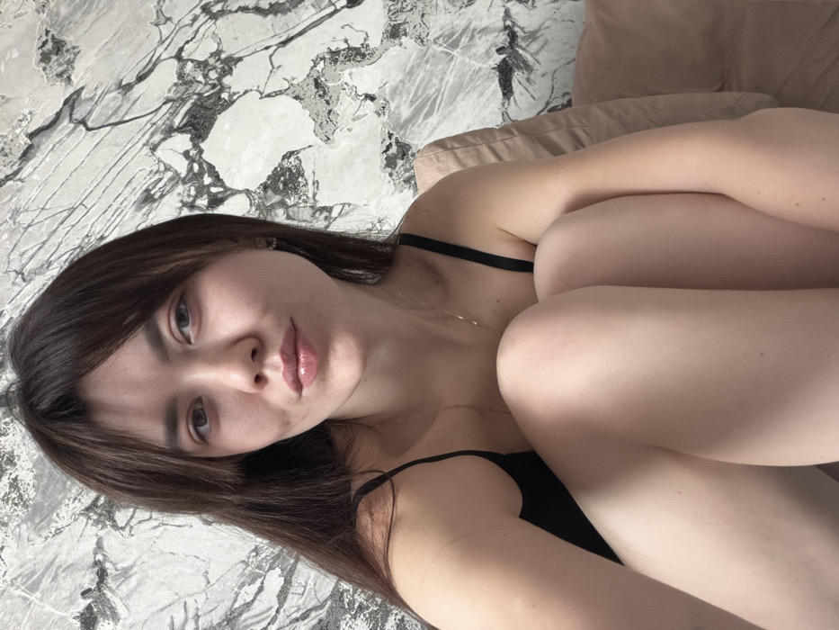 A young adult female is depicted in a close-up, indoor shot. She has long, dark brown hair with bangs, light eyes, and fair skin, with a neutral expression and slightly pursed lips, looking directly at the viewer. She wears a black spaghetti-strap top and a thin gold-colored necklace. Her body is positioned with knees drawn up, visible in the lower frame, indicating a seated or reclining posture. A light-colored, possibly beige, cushion or pillow is visible behind her right shoulder. The background features a wall covered in a light-toned material with a prominent white and grey marble pattern, exhibiting dark, irregular veining. The specific location within the city of Chaikovsky, Russia, cannot be ascertained from the visual information provided in the image.