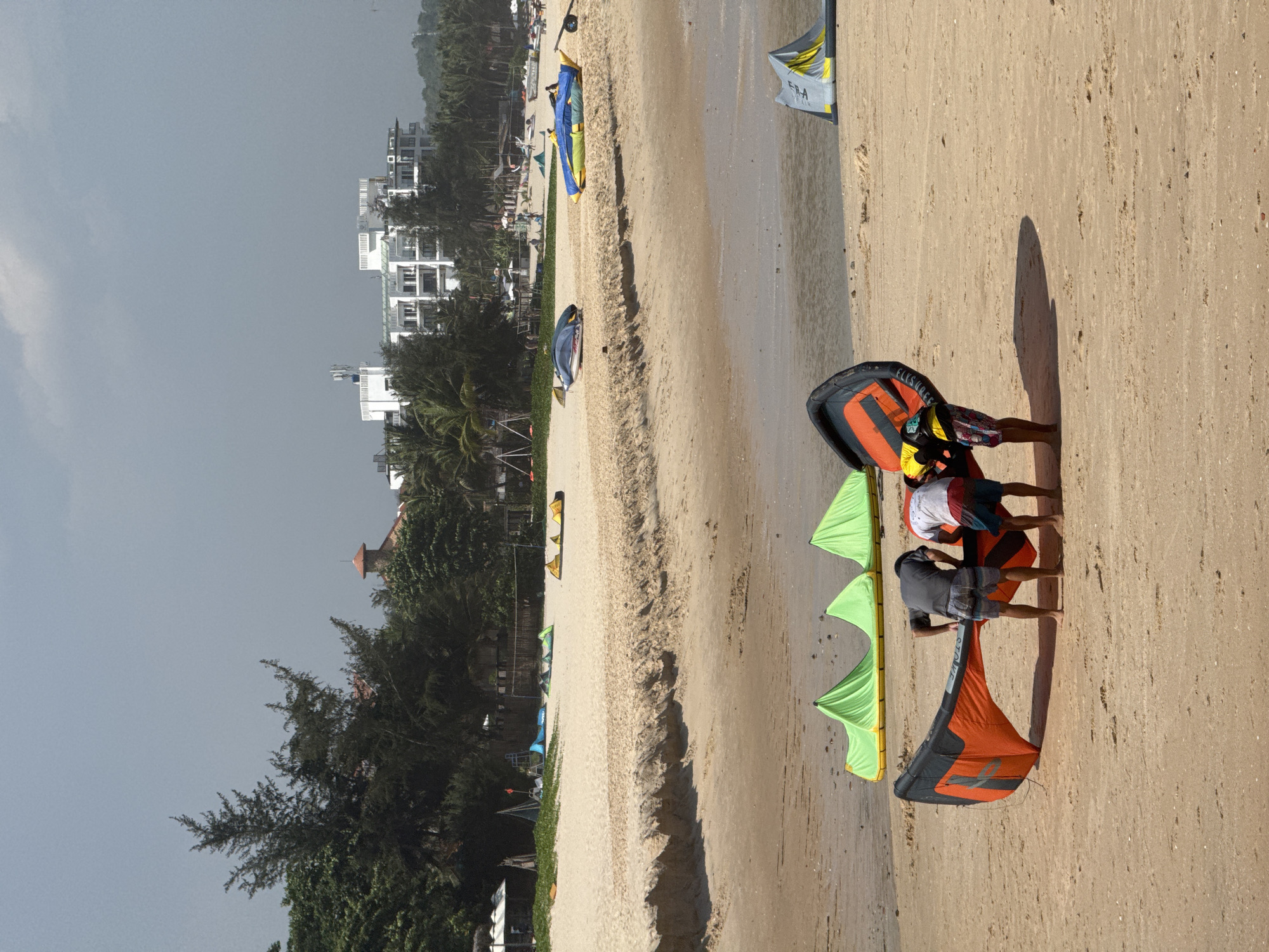 A sunny day at Mũi Né, Vietnam, shows a sandy beach with several kitesurfing kites laid out on the sand. In the foreground, three individuals are preparing an orange and black kite, two are bending over it and one is standing nearby. In the middle ground, several green kites are spread out on the sand. In the background, a line of palm trees and lush vegetation separates the beach from a cluster of modern white buildings. The sky is overcast.