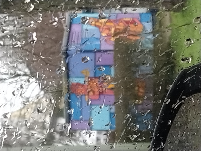 A rectangular display featuring a colorful geometric design with orange figures is visible through a rain-streaked window. The background includes trees. The dominant colors on the display are light blue, purple, and orange. A partial view of a car door is seen on the right side of the frame. The raindrops distort the image, creating a blurry effect.