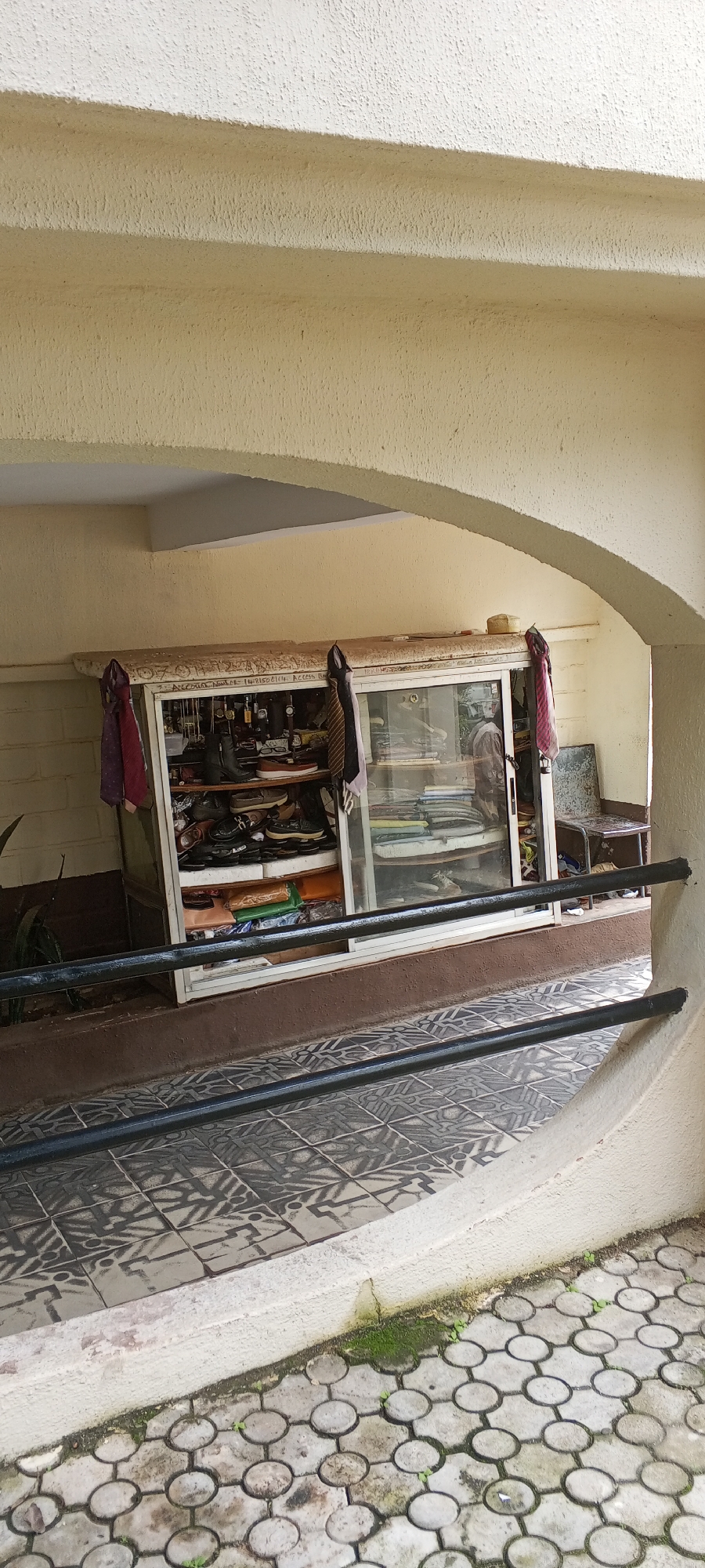 The image shows a display cabinet filled with various items, including shoes, watches, and folded clothing. The cabinet is situated outdoors, possibly in a shop or market stall, under an archway. The floor in front of the cabinet is tiled with a patterned design. In the background, there is a brick wall and a metal chair. The environment appears to be a street or an outdoor commercial area. The writing on the top of the cabinet suggests it may be a business or a vendor. There are no people visible in the image. The overall impression is of a modest retail display in an urban setting. The location is identified as the city of Jos, Nigeria, though no specific landmarks or context are provided in the image itself to confirm this.