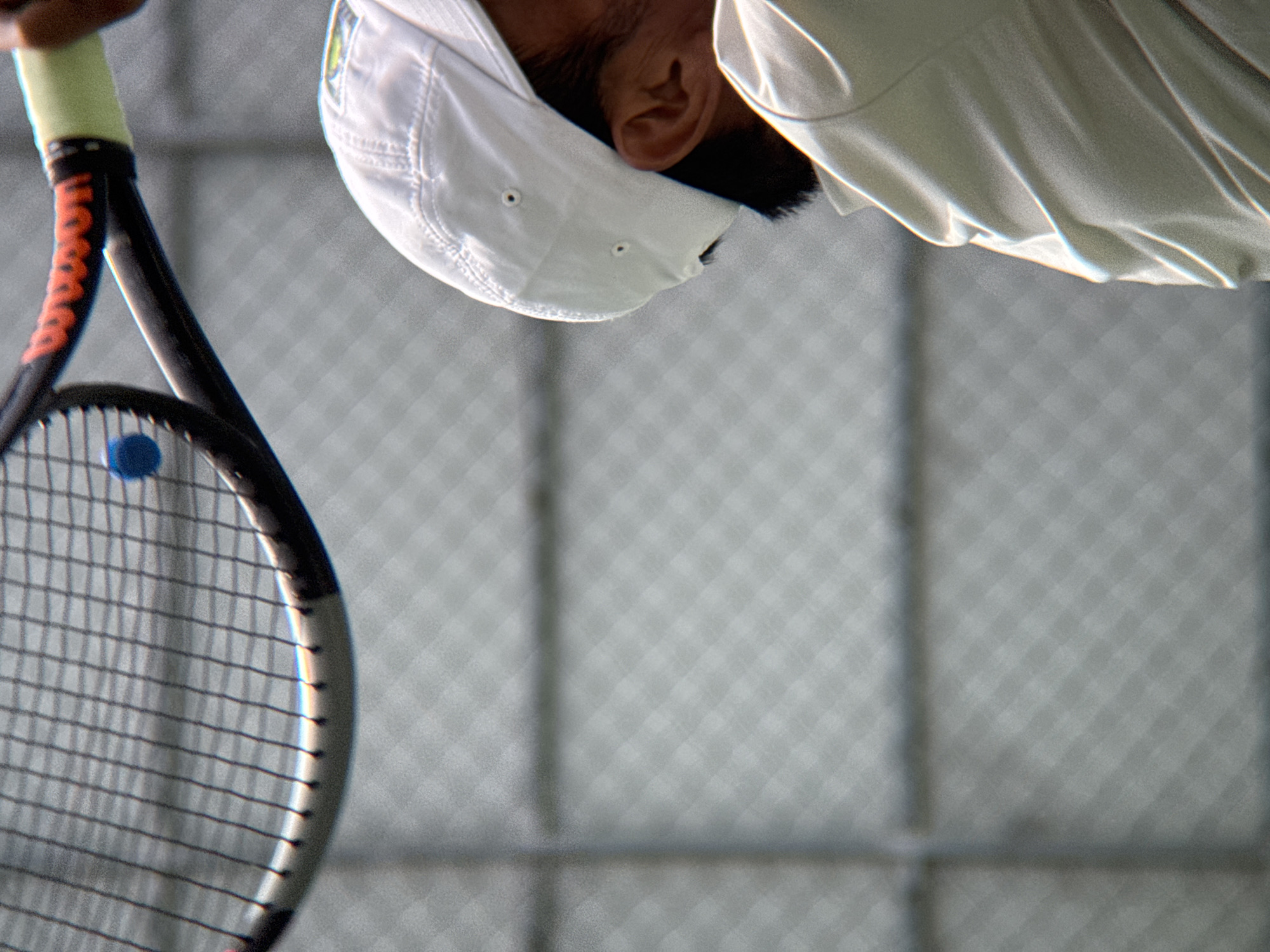 In Nha Trang, Vietnam, a tennis player is captured in motion, mid-swing, as their racket connects with a blue tennis ball. The player, wearing a white cap and light-colored attire, is positioned in the foreground, with their head and upper body visible. The tennis racket, a Wilson brand, is in the process of hitting the ball, which is suspended in the air just above the strings. The background features a chain-link fence, suggesting an outdoor tennis court environment. The lighting indicates an outdoor setting, likely during the daytime.