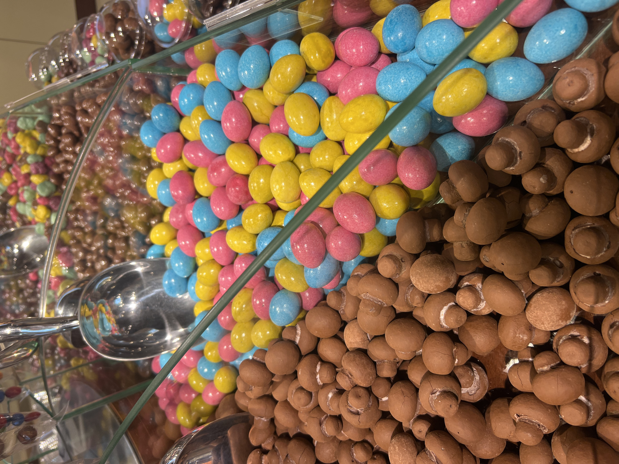 The image shows a close-up, eye-level view of a confectionery display case filled with various sweets. In the foreground, to the left, are numerous chocolate-covered candies shaped like mushrooms, predominantly brown in color. To the right of the mushroom candies, and filling the majority of the frame, are brightly colored, oval-shaped candies in shades of pink, yellow, and blue. These are arranged in a pile, with a metallic scoop partially visible in the upper left quadrant, suggesting they are intended for self-service. The display case appears to be made of glass, with a green border visible diagonally across the frame. In the background, more candies in various containers are visible, some appearing to be chocolate balls in shades of brown and other colors. The lighting is bright, creating reflections on the glass and the candies. There are no people or visible signs indicating a specific location within Odesa, Ukraine. The scene depicts a retail environment focused on confectionery.