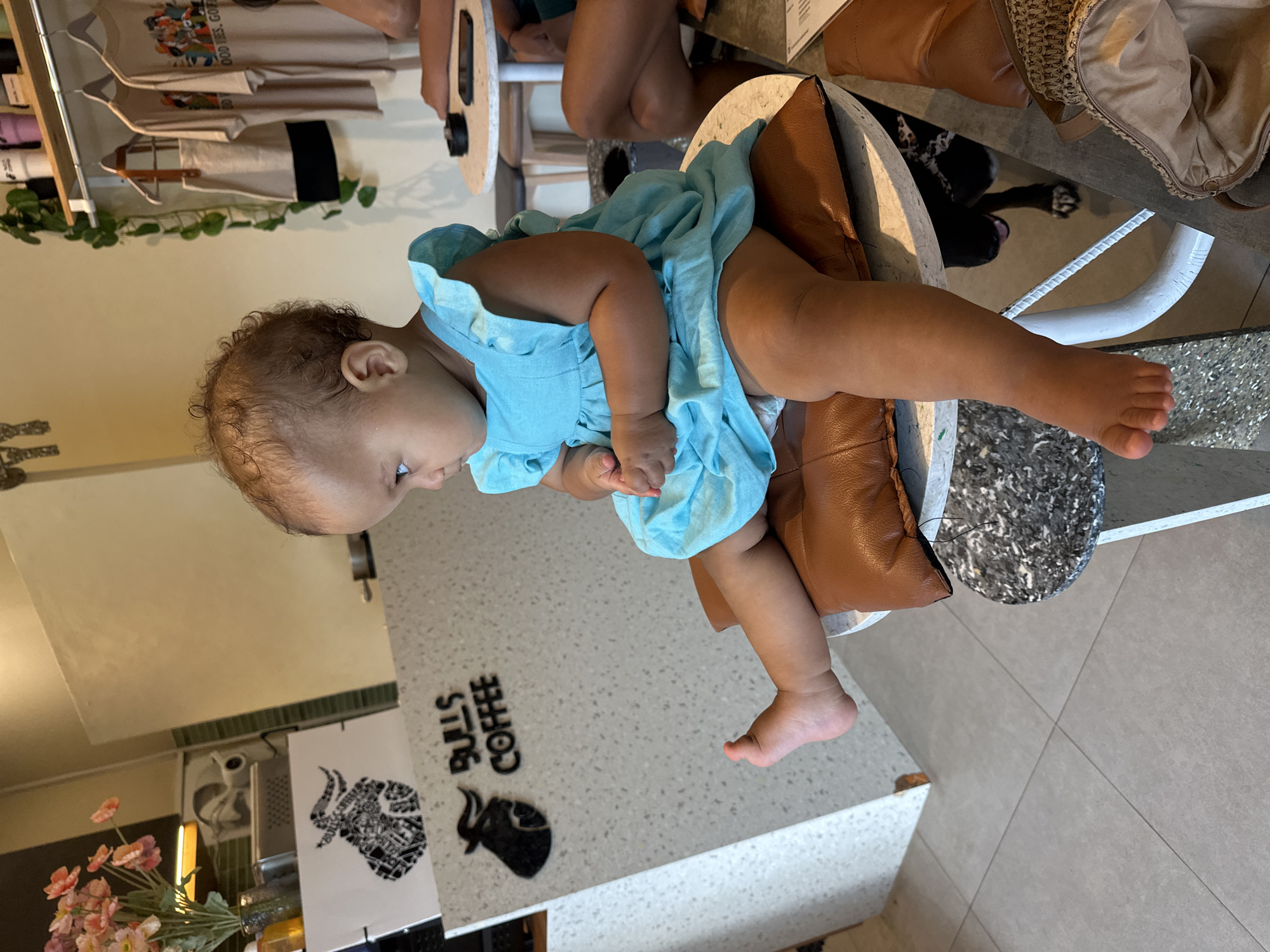 A light-skinned baby girl is sitting on a brown cushion on a speckled table. She is wearing a turquoise dress and is looking to the right. The background shows a cafe with a counter displaying various items. A black dog with a white collar is partially visible under the table. The cafe appears to be in North Kuta, Indonesia.
