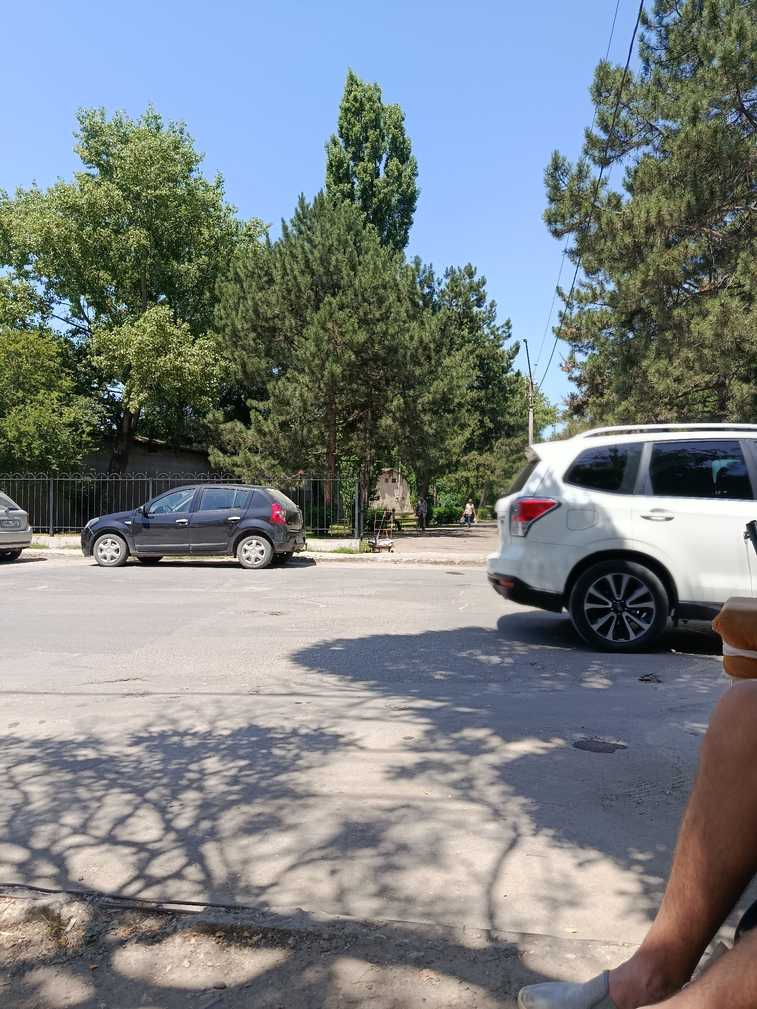 In the city of Bălți, Moldova, on a clear, sunny day, a dark-colored hatchback car is parked on the side of a paved street. The car, appearing somewhat dusty, has its side facing the viewer, and a black metal fence is visible behind it. To the right of the hatchback, a white SUV is parked, also on the side of the street, with its rear passenger side facing the camera. The background features numerous tall, green trees and a bright blue sky. In the distance, on the left side of the frame, several people are walking, and some are seated near a building. The foreground shows the street surface with shadows of tree branches cast across it. A person's legs, wearing light-colored slip-on shoes, are visible in the lower right corner of the image, indicating the photographer's presence.