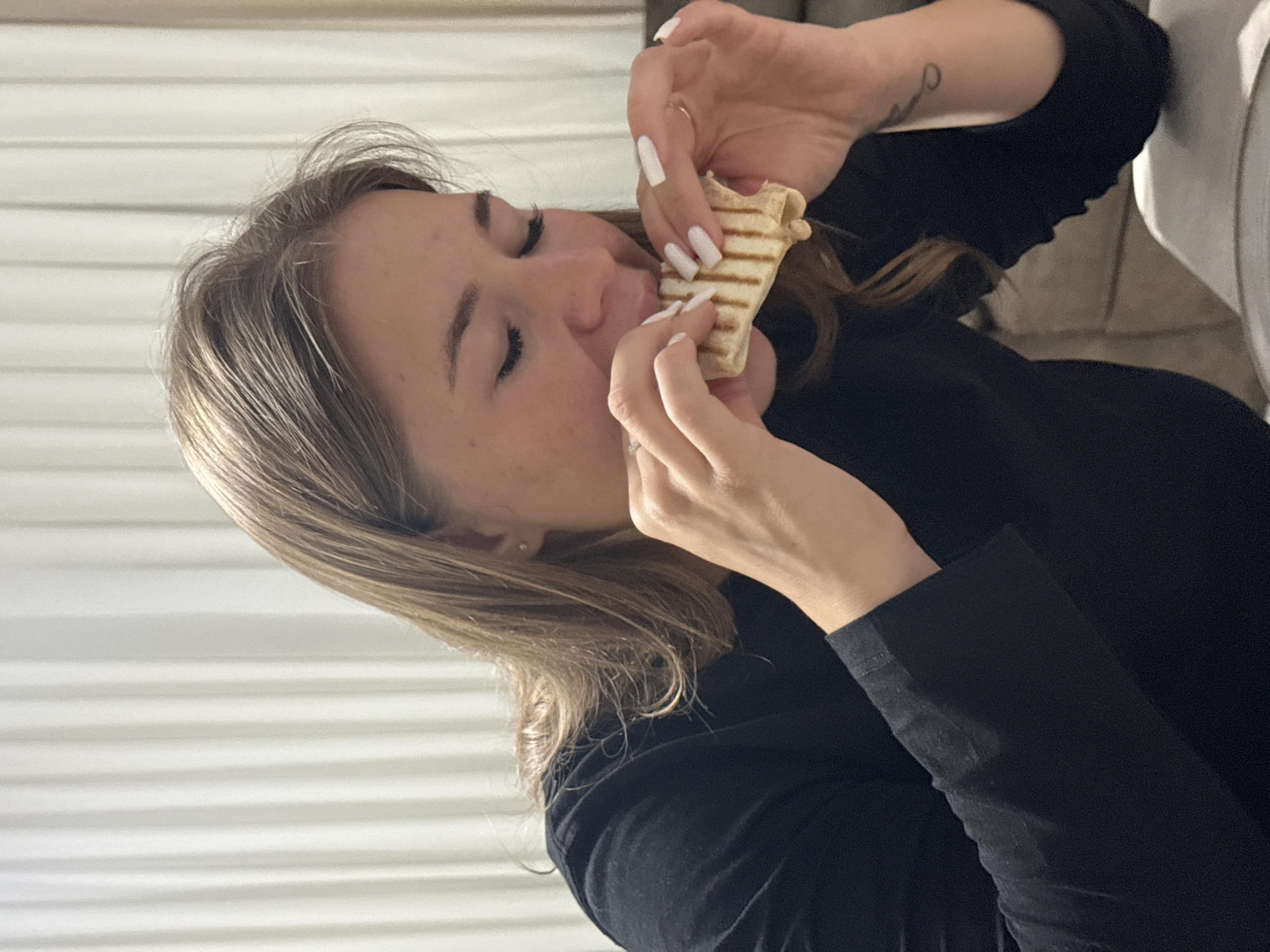 The image depicts a female subject in an indoor setting, actively consuming a grilled sandwich. The subject, with light brown hair, is wearing a black long-sleeved garment. She holds the sandwich with both hands, which display white, square-tipped fingernails. Her eyes are closed or cast downwards as she bites into the food. A silver-toned ring is visible on her left ring finger, and a gold-toned ring is on her right ring finger. A small, dark tattoo is present on the inside of her right forearm. A small stud earring is visible in her right ear. The sandwich has distinct grill marks. The background features white vertical blinds or ribbed curtains on the left and center, with a light-colored upholstered sofa or armchair partially visible on the right. A white surface, possibly a plate or table, is in the lower right foreground. There are no identifiable visual markers within the image to confirm a specific location in the city of Budva, Montenegro.