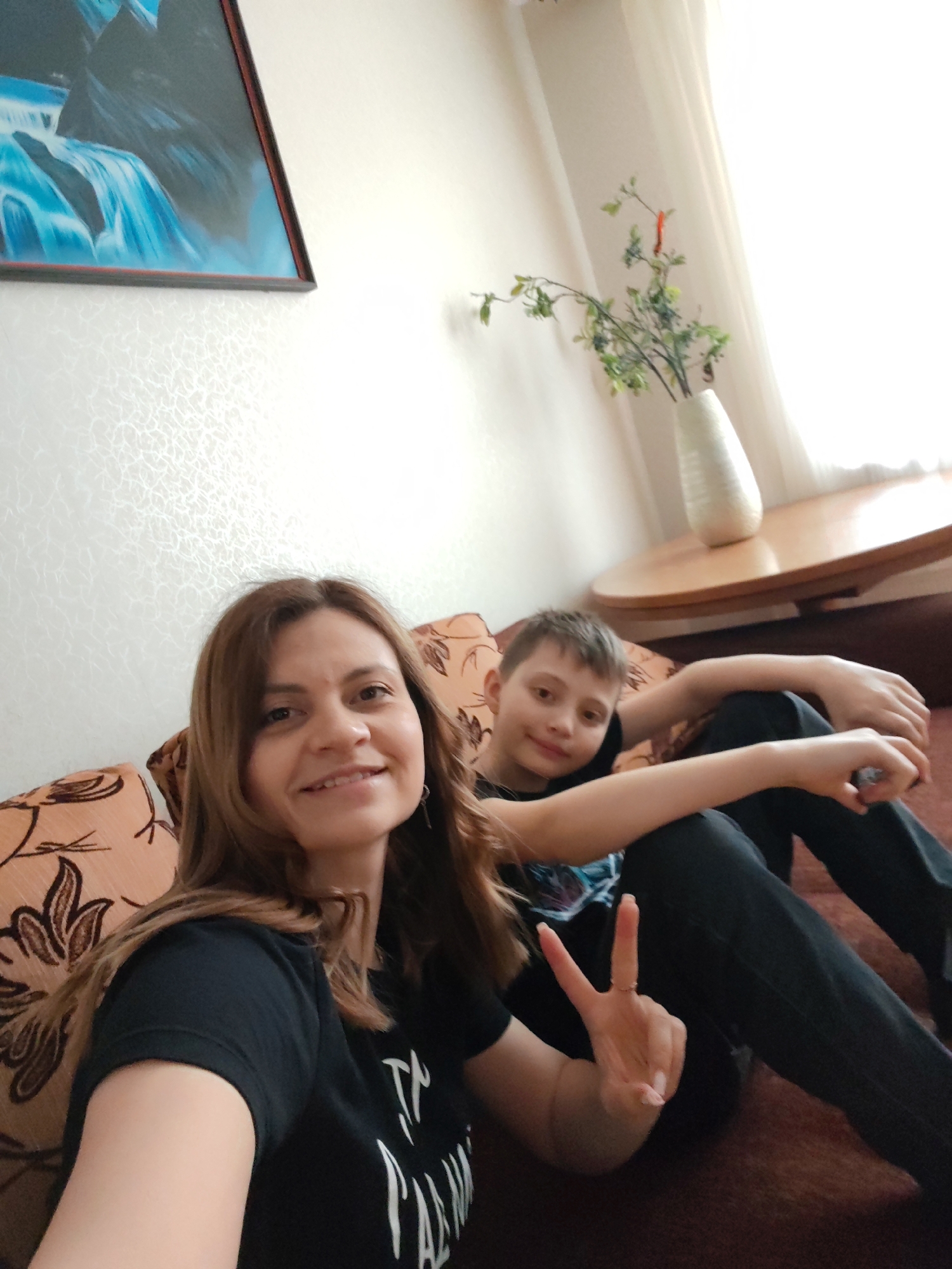 A medium-shot photograph depicts a woman and a boy, presumably indoors, seated on a couch. The woman, positioned in the foreground, is smiling and displaying a peace sign with her right hand. She is wearing a black t-shirt with white text. Behind her to the right sits a boy with his arms resting on his lap. He is wearing a dark t-shirt. The couch is upholstered with fabric featuring a brown floral pattern. Behind them, against a textured wall, is a framed picture with a blue waterfall scene. To the right, near a window, is a round wooden table with a white vase containing green stems. The location is indicated to be in the city of Unknown, Unknown.