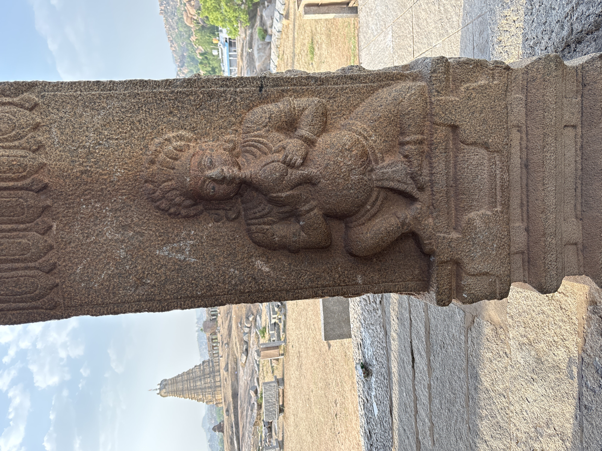 The image shows a close-up of a carved stone pillar in Hampi, India. The carving depicts a figure, possibly a deity or mythical being, in a reclining or resting position. The figure has detailed features, including facial expressions, intricate hair or headwear, and a rotund body. The stone itself has a rough, textured surface. In the background, there are glimpses of the Hampi landscape, including other stone structures, a temple tower, and open ground. The sky is visible in the upper background. The overall scene conveys a sense of ancient artistry and the historical significance of Hampi.