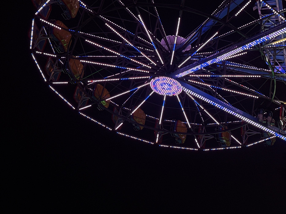 A large Ferris wheel, illuminated with numerous lights, is shown against a dark night sky. The wheel's metallic structure is outlined by small, predominantly white and blue LED lights. The central hub features concentric rings of white, blue, and magenta lights. Multiple passenger gondolas, colored orange, yellow, and green, hang from the wheel's perimeter; no individuals are clearly discernible within them. The image depicts a nighttime scene in Mexico City, Mexico.