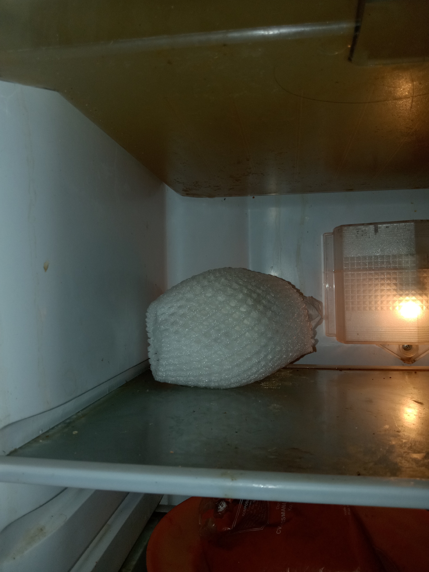 This is an interior shot of a refrigerator, likely in a domestic setting. The refrigerator appears to be at least partially empty, with a single, large, white, textured object prominently placed on the middle shelf. This object resembles a packaged food item, possibly frozen, due to its round, somewhat compressed shape and the bubble wrap-like texture on its surface. The interior of the refrigerator shows signs of wear and some discoloration on the walls and ceiling, suggesting it is not new. The light fixture on the right side of the interior is illuminated, casting a warm glow. Below the middle shelf, partially visible, is a dark red or brown plastic bag with some visible text in what appears to be Indonesian. The overall impression is of a refrigerator in use, containing a single, wrapped item. Without further context, the specific location in Jumput, Indonesia cannot be determined, but the presence of Indonesian text on the bag suggests a connection to the region.