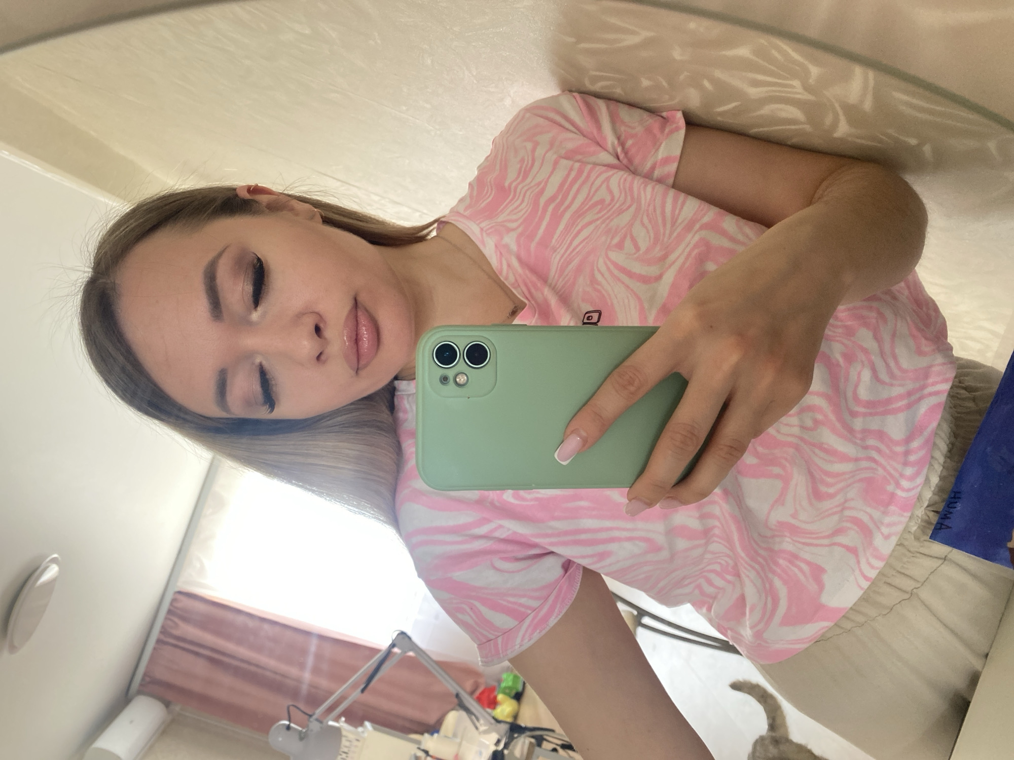 A young woman is taking a selfie in a mirror. She is wearing a pink and white patterned t-shirt and light-colored shorts. She is holding a light green smartphone in her right hand. The background shows a room with a desk lamp, various objects on the desk, and a partially visible window. The scene appears to be indoors, possibly a bedroom or study. There is a cat's tail visible near the bottom edge of the frame. There is no indication of the location being Izhevsk, Russia, or any other city.
