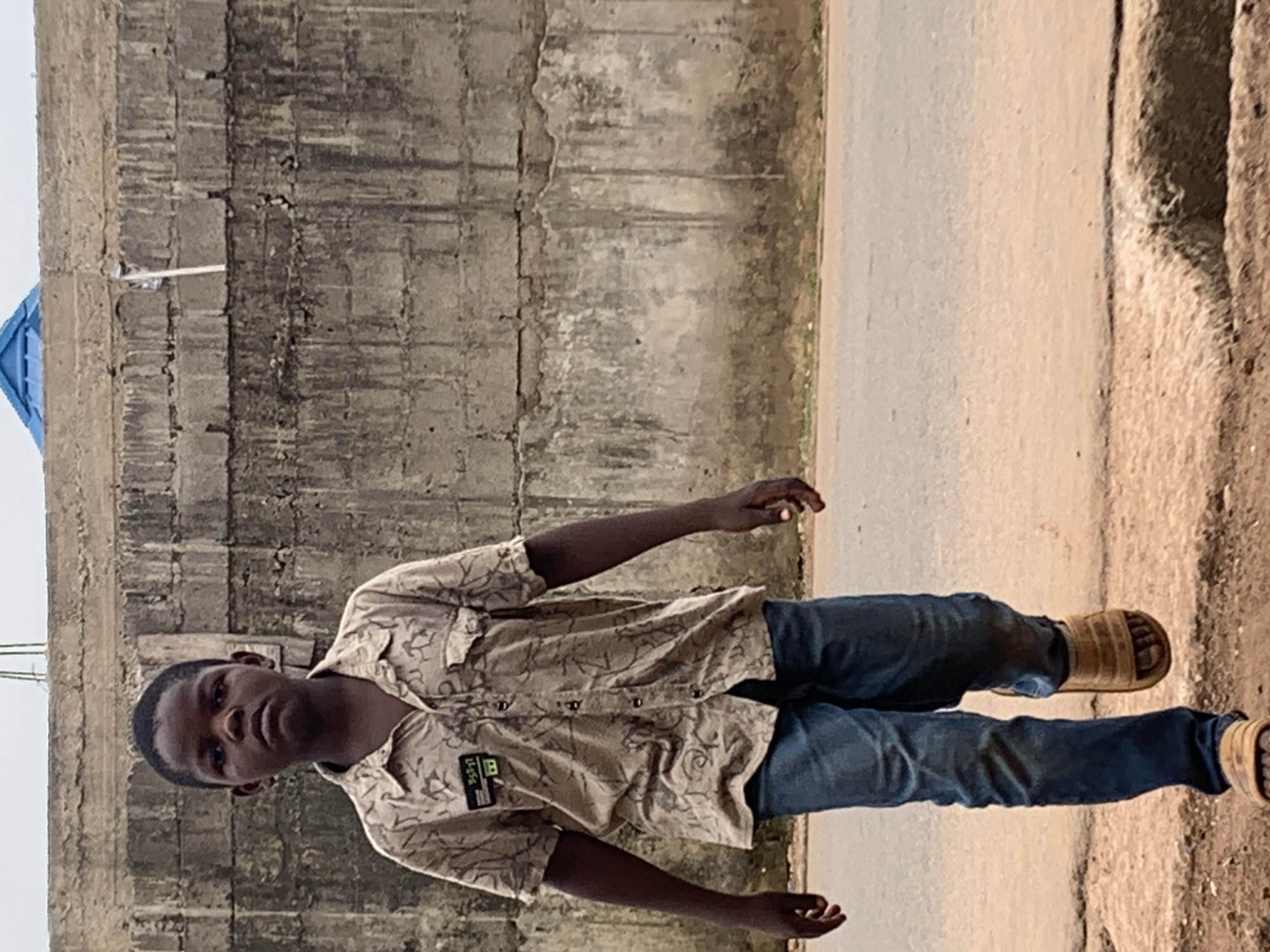 A young man stands on a paved surface next to a concrete wall in Akwanga, Nigeria. He wears a patterned tan shirt, blue jeans, and brown sandals. The wall appears weathered, and a small white fixture is attached to it. The paved surface is bordered by rougher terrain.