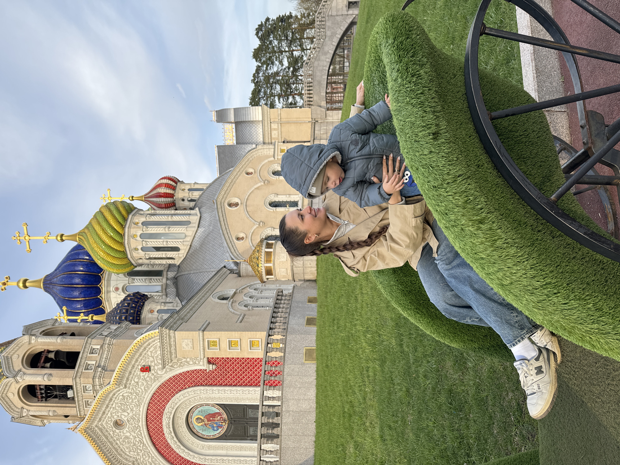 A woman and child are positioned in front of the Cathedral of the Russian Armed Forces in Moscow, Russia. The woman is fair-skinned with her dark hair in a braid, wearing a beige jacket and blue jeans with white sneakers. She is interacting with a young child wearing a blue jacket, who is in her arms. They are situated on a structure covered in artificial green turf, with a decorative black metal wheel adjacent to them. The Cathedral, a large religious building with multiple colorful domes and intricate architectural details, is in the background. A green lawn is visible around the building. The sky is partly cloudy.