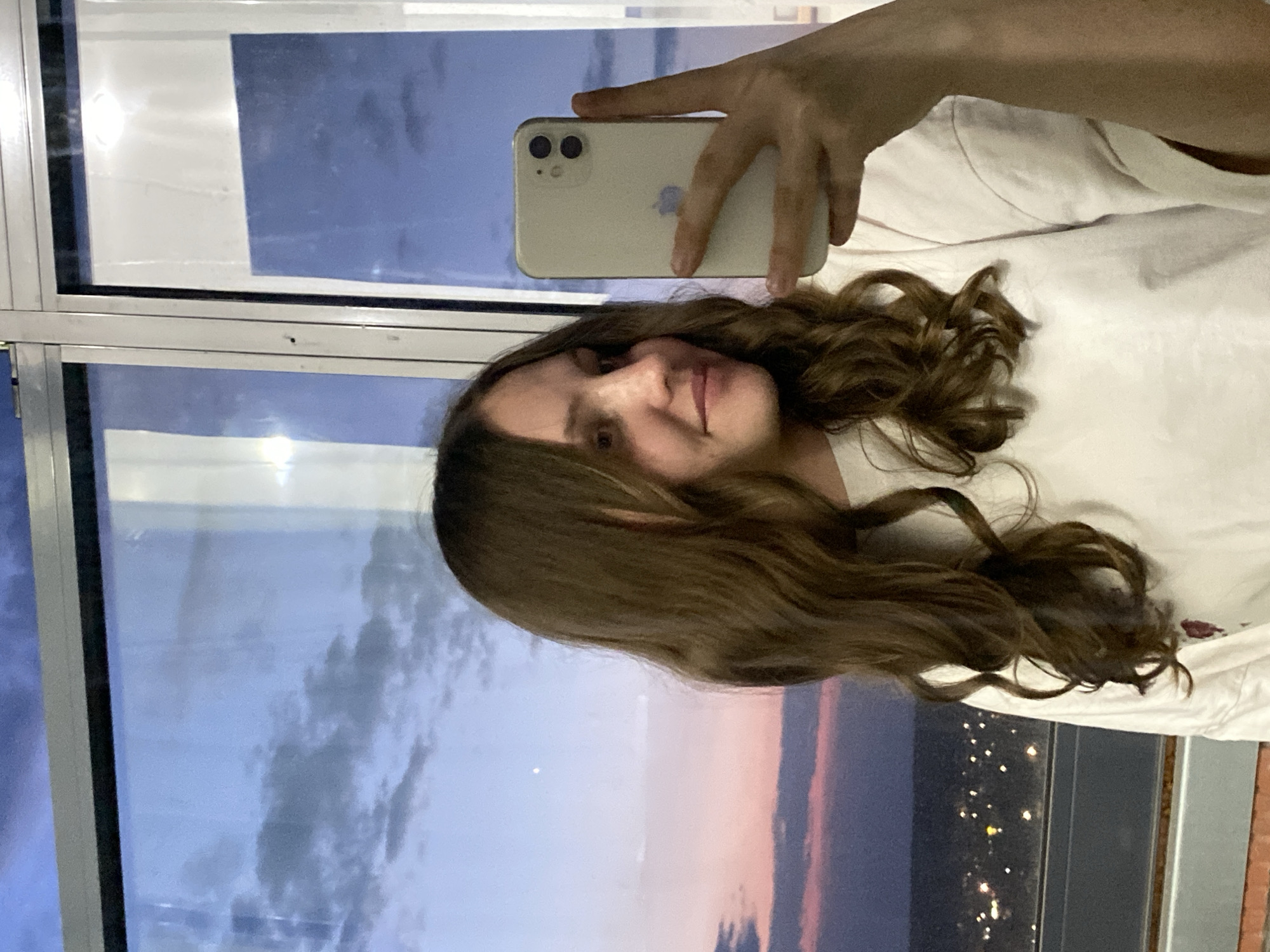 A woman with brown, wavy hair is holding a smartphone, presumably taking a selfie. She is wearing a white shirt. The background features a window with a view. The reflection in the glass shows a sunset sky with pink and purple hues transitioning to a night scene with city lights, indicating an urban environment. Given the prompt, it is possible the location is Izhevsk, Russia, based on the cityscape visible in the reflection.