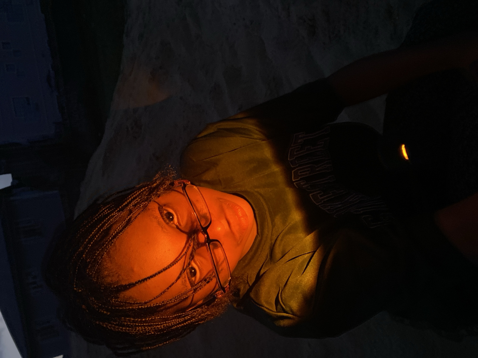 The image displays a single individual in Iranla, Nigeria, under low-light conditions, illuminated by an intense, warm orange-yellow light source from below. The subject, appearing to be a young person with dark skin and braided hair, wears clear-framed eyeglasses and a dark-colored t-shirt with white lettering on the chest, partially legible as "STREET." The individual looks directly into the camera with a serious expression, their head tilted slightly upwards. The warm light prominently highlights the face, particularly the forehead, eyes, and mouth, while casting deep shadows on the upper head and sides. A distinct, bright orange glow is visible on the lower front of the subject's shirt. The background is dark and out of focus, revealing a slightly elevated, sandy or dusty ground surface to the left and faint silhouettes of dark buildings or structures against a dark sky. The scene suggests an outdoor setting during twilight or night.