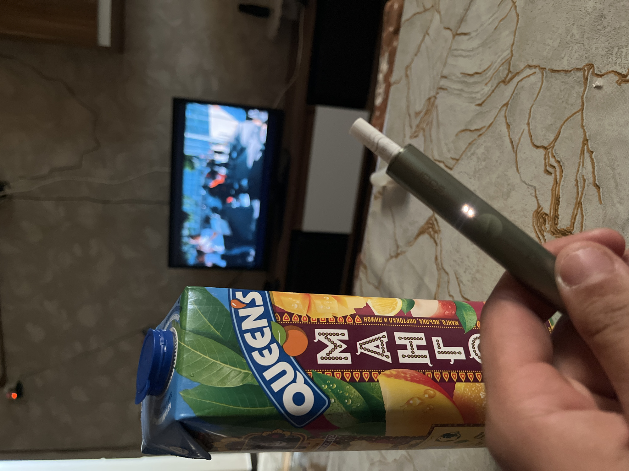 A hand holds an electronic cigarette over a carton of "Queens" brand mango, apple, orange and lemon juice. In the background is a television. The location is likely indoors. The location in the city of Simeonovgrad, Bulgaria cannot be determined.