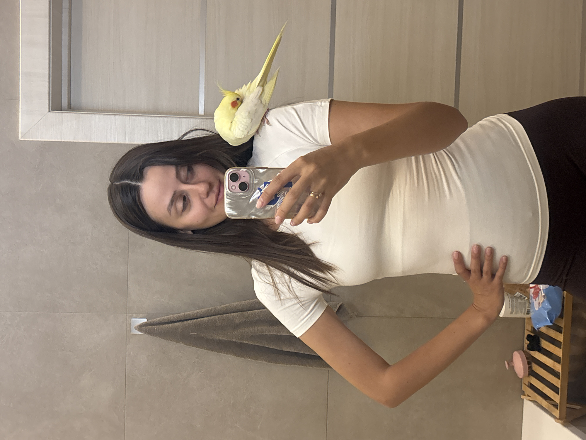 In Krasnoyarsk, Russia, a woman takes a mirror selfie in a bathroom. She is wearing a white t-shirt and dark pants. A yellow cockatiel is perched on her left shoulder, pecking at its wing. The woman is holding a smartphone in her right hand, using it to capture the reflection. The bathroom features tiled walls, a towel hanging on a hook, and a wooden shelf with various items on it. A door is visible in the background.