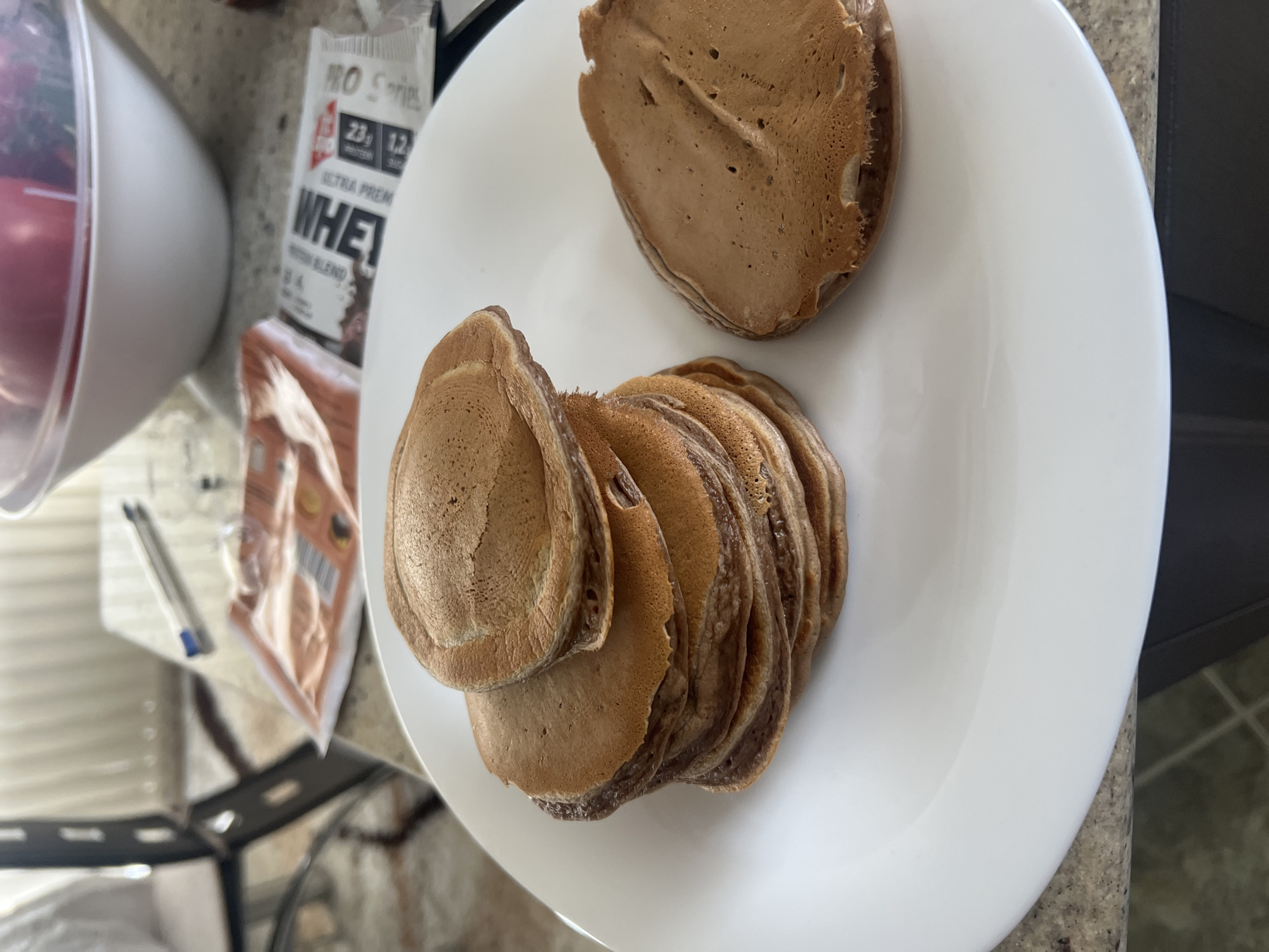 The image shows a plate of pancakes on a kitchen counter. There is a stack of multiple small pancakes in the lower center of the plate, and a larger pancake resting above the stack. A container and a package of protein whey mix are visible next to the plate. A portion of a chair is seen in the lower left of the image. The location is Unknown, Unknown.