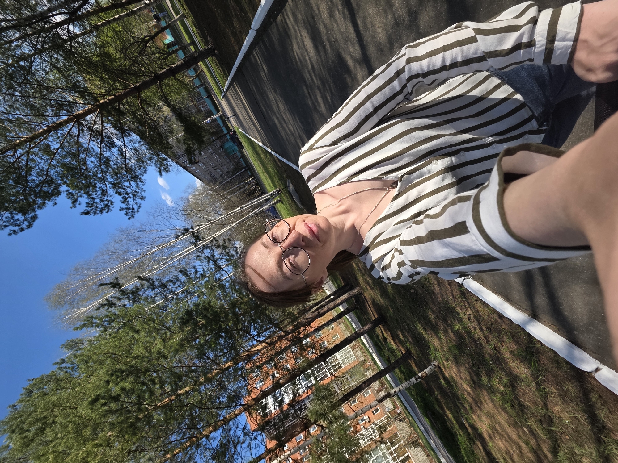 The image shows a woman taking a selfie outdoors in the city of Chaikovsky, Russia. She is wearing glasses and a horizontally striped shirt. Behind her are trees, a building with red bricks, and a clear blue sky. The scene is well-lit, indicating it's daytime. The woman is likely sitting or standing near a road or pathway. The photo is taken at an angle, with the camera held above the subject.