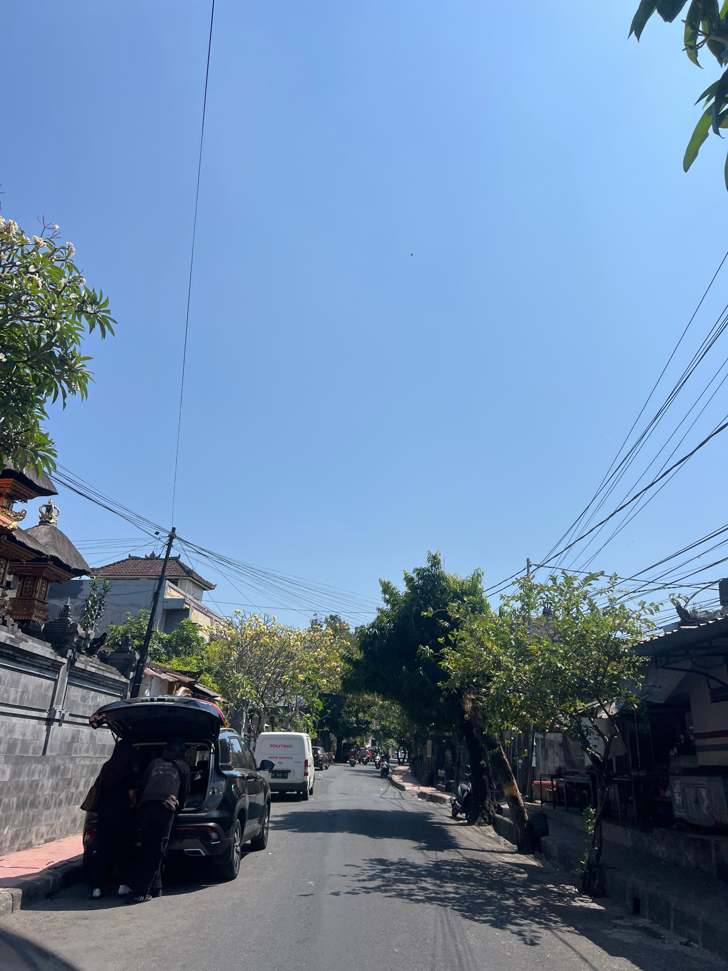 This image depicts a street scene in Denpasar, Indonesia, likely on a sunny day given the clear blue sky and strong shadows. The perspective is from a low angle, looking down a paved street lined with buildings and trees.
On the left side of the street, there is a traditional-looking stone wall with ornate architectural details, possibly part of a temple or a distinctive residential property. Behind the wall, buildings with tiled roofs are visible, some featuring elaborate decorative elements. A lush green tree with white flowers is prominent in the foreground on the left.
In the middle of the street, a dark-colored SUV is parked, its trunk open. Two individuals, dressed in dark clothing, appear to be engaged in loading or unloading items from the vehicle. A white van with the word "POLYTRON" and "HOME SERVICE" written on its side is also visible further down the street.
The right side of the street features a row of low-rise buildings with awnings, suggesting shops or residences. Trees with dense green foliage and some trees with yellow flowers provide shade along the sidewalk. Various electrical wires crisscross overhead, supported by utility poles. Several motorcycles are parked along the street and a few are visible in motion further down the road, indicating active local traffic. The street surface is asphalt, with visible markings. The overall impression is a lively, everyday scene in a tropical urban environment.