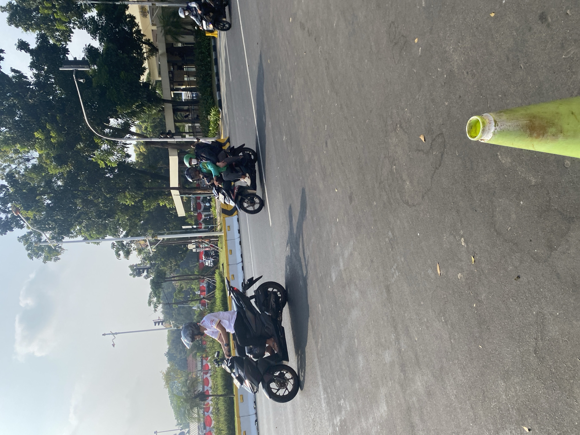 This is a photograph taken from a slightly elevated, eye-level perspective on a paved road in Central Jakarta, Indonesia. The foreground shows a grey asphalt road with some scattered leaves and debris. A bright green traffic cone with a white band is visible in the lower right corner, angled towards the left.
In the middle ground, a black motorcycle is being ridden by a person wearing a white t-shirt, black pants, and a dark helmet. The rider's left hand is on the handlebar, and their right hand is visible. The motorcycle has a dark frame and silver wheels.
Further back on the road, another motorcycle carries two people. The rider is wearing a green jacket and a dark helmet, and the passenger behind them is wearing a dark jacket and a green helmet. Both appear to be wearing face masks.
The background features a row of trees with dense green foliage, some of which are illuminated by sunlight. Behind the trees, a low-rise building with a white facade and a dark-tiled roof is visible. There are also streetlights and traffic signals visible along the roadside. In the far background, there are hints of buildings and more trees, suggesting an urban environment. The sky is a light blue with some scattered white clouds, and the overall lighting indicates it is daytime.