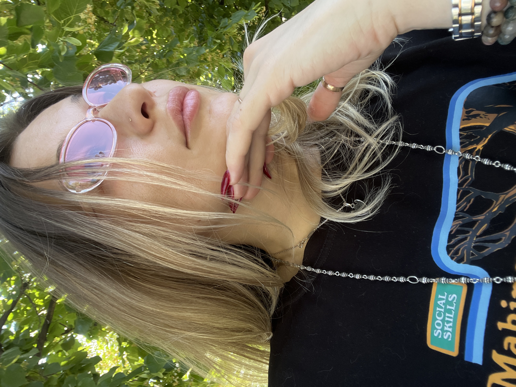 A woman with blonde hair and pink-tinted sunglasses is seen from the chest up, resting her chin on her hand. She is wearing a black t-shirt with a graphic design and jewelry. The background consists of green foliage, suggesting an outdoor setting. The lighting indicates it is daytime, possibly sunny given the brightness and shadows.