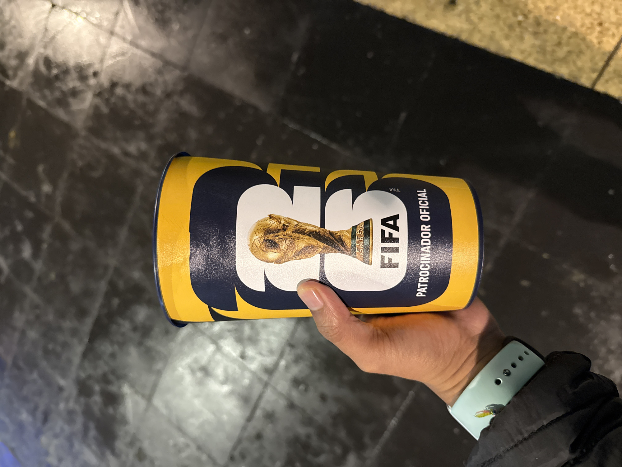A hand wearing a light blue smartwatch and a black jacket sleeve holds a tall, yellow and navy blue disposable cup. The cup features a graphic of the FIFA World Cup trophy in gold, with the FIFA logo and the words "PATROCINADOR OFICIAL" printed below it in white. The cup is positioned in front of a dark, textured surface that appears to be a tiled floor, possibly in a public space or venue. The lighting suggests an indoor or dimly lit outdoor environment.