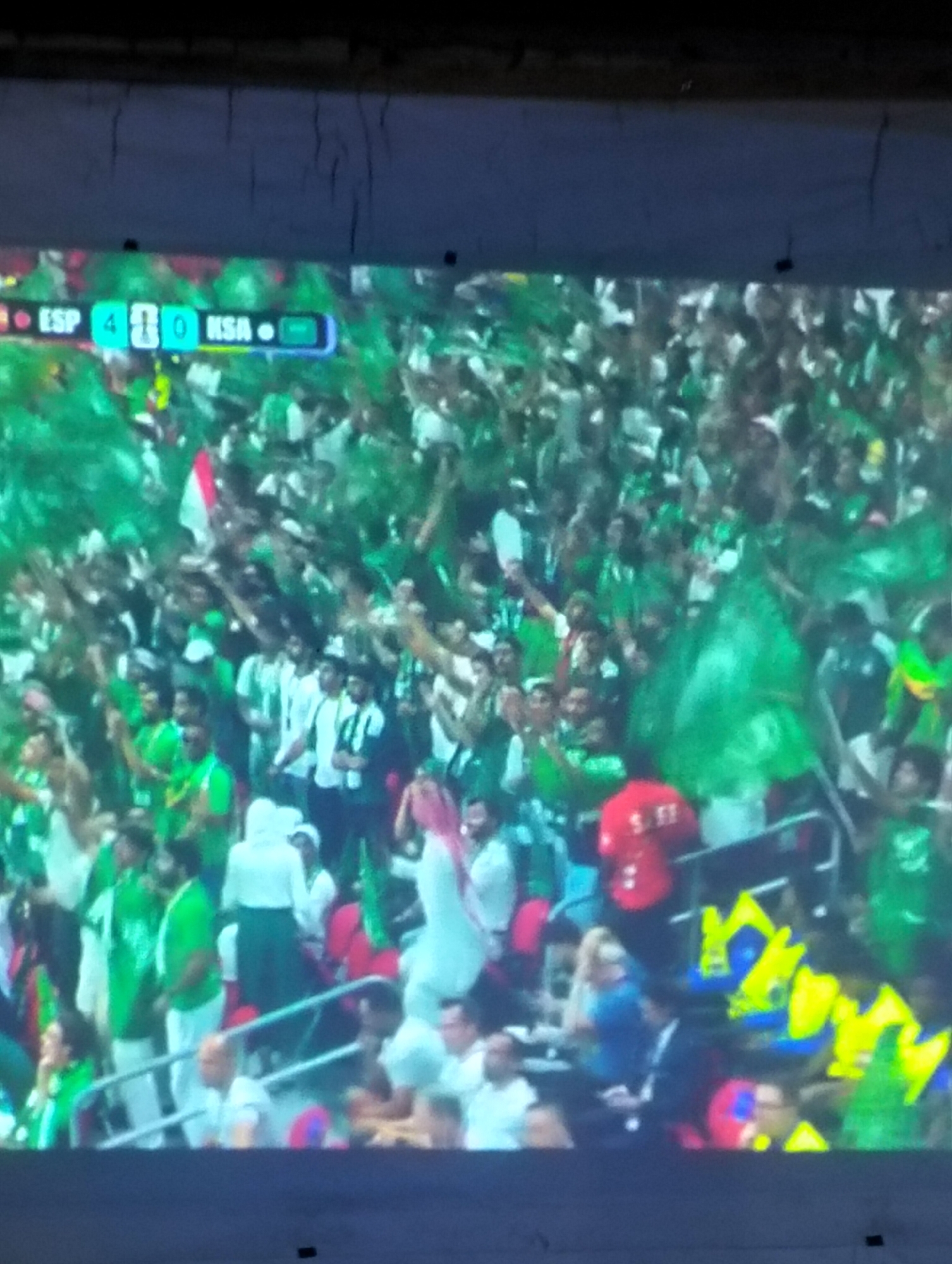 The image shows a crowd of people at a sporting event, likely a soccer match, given the score display at the top left. The score reads "ESP 4 - 0 KSA," indicating a game between Spain (ESP) and Saudi Arabia (KSA) with Spain leading 4-0. The crowd is largely dressed in green, with some individuals wearing white or red. Many people are holding green flags and appear to be cheering, suggesting a lively and enthusiastic atmosphere. The setting appears to be a stadium, with tiered seating visible in the background. The lighting suggests it is daytime. There is no visible text other than the score. The overall impression is of a large gathering of fans supporting their team.
