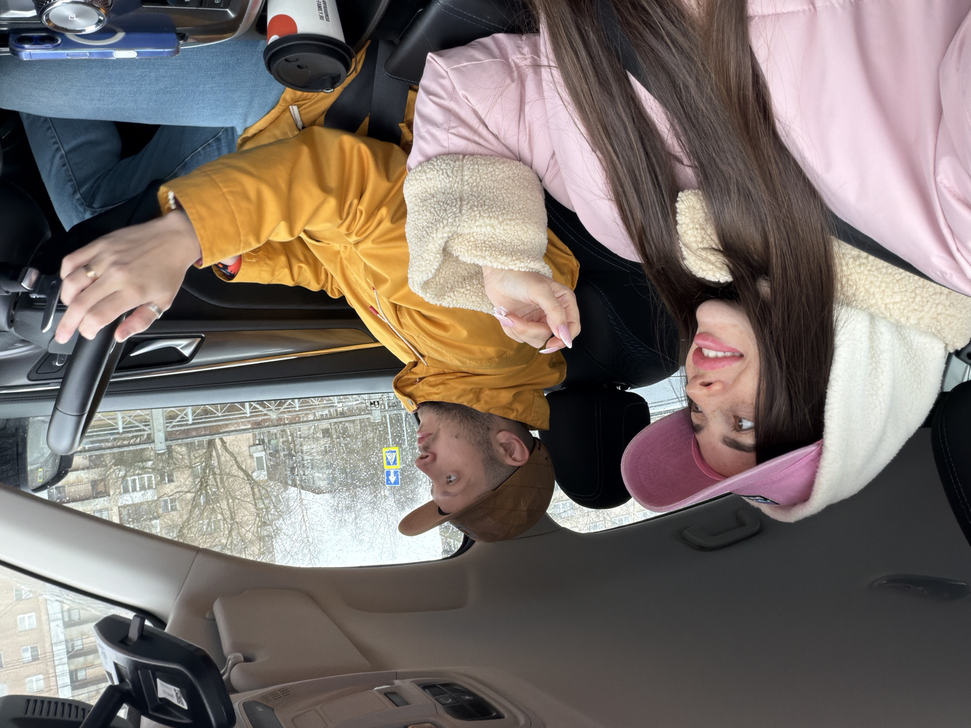 A couple is in a car. The man, wearing a brown cap and yellow jacket, is driving. The woman, wearing a pink jacket and matching baseball cap with a white hood, is in the passenger seat. A coffee cup is on the console. Outside, it is cloudy and possibly raining, and buildings characteristic of Saint Petersburg, Russia, are visible through the windshield.
