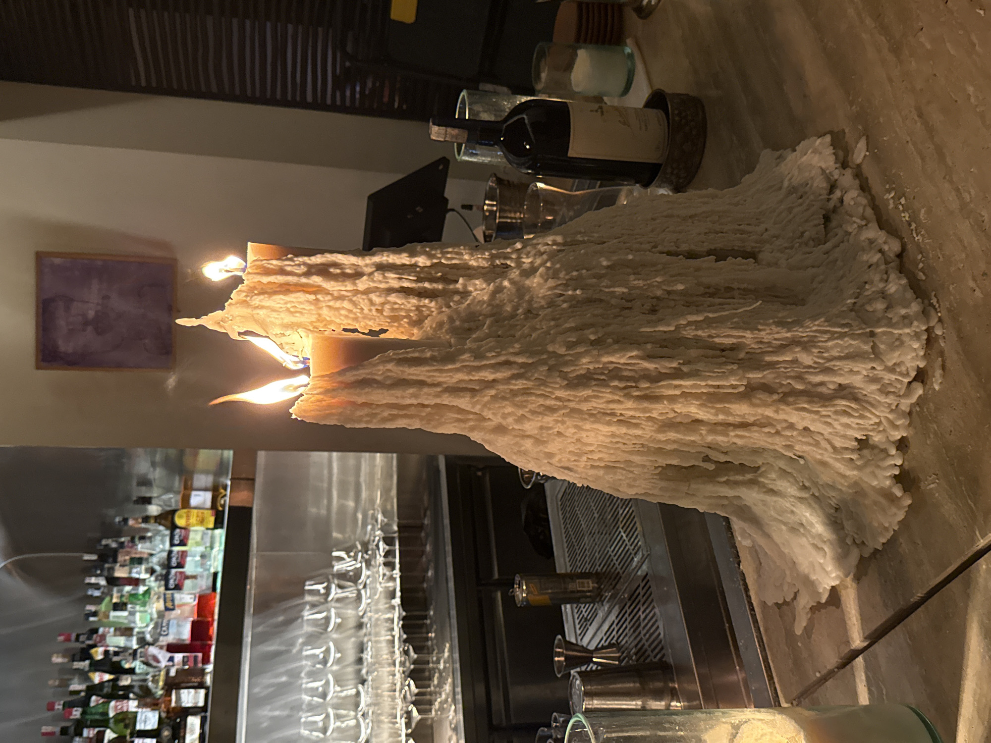The image captures a dimly lit interior scene, likely a bar or restaurant in Kuta Selatan, Indonesia, illuminated primarily by a massive, multi-wick candle. The central subject is this large candle, which has accumulated an enormous amount of melted and re-solidified wax, creating a highly textured, irregular, and flowing form that dominates the foreground and midground. Two distinct flames are visible, casting warm, orange light that highlights the craggy surfaces of the hardened wax.
In the background on the left, a bar area is visible, stocked with numerous bottles of various spirits and liqueurs, neatly arranged on shelves. Below these bottles, rows of clean, inverted glassware, possibly wine glasses, are placed on a stainless steel surface, alongside other bar tools like jiggers and shakers. On the wall further back, a framed picture with a dark, indistinct, purplish artwork hangs.
To the right of the candle, a dark bottle of wine with a light-colored label stands, accompanied by more glassware. The overall lighting suggests evening or nighttime, creating an intimate and warm ambiance. The significant wax buildup on the candle indicates it has been burning for an extended period, serving as a prominent decorative element. No specific human activity is depicted, and no readable text is visible on any labels or signs.