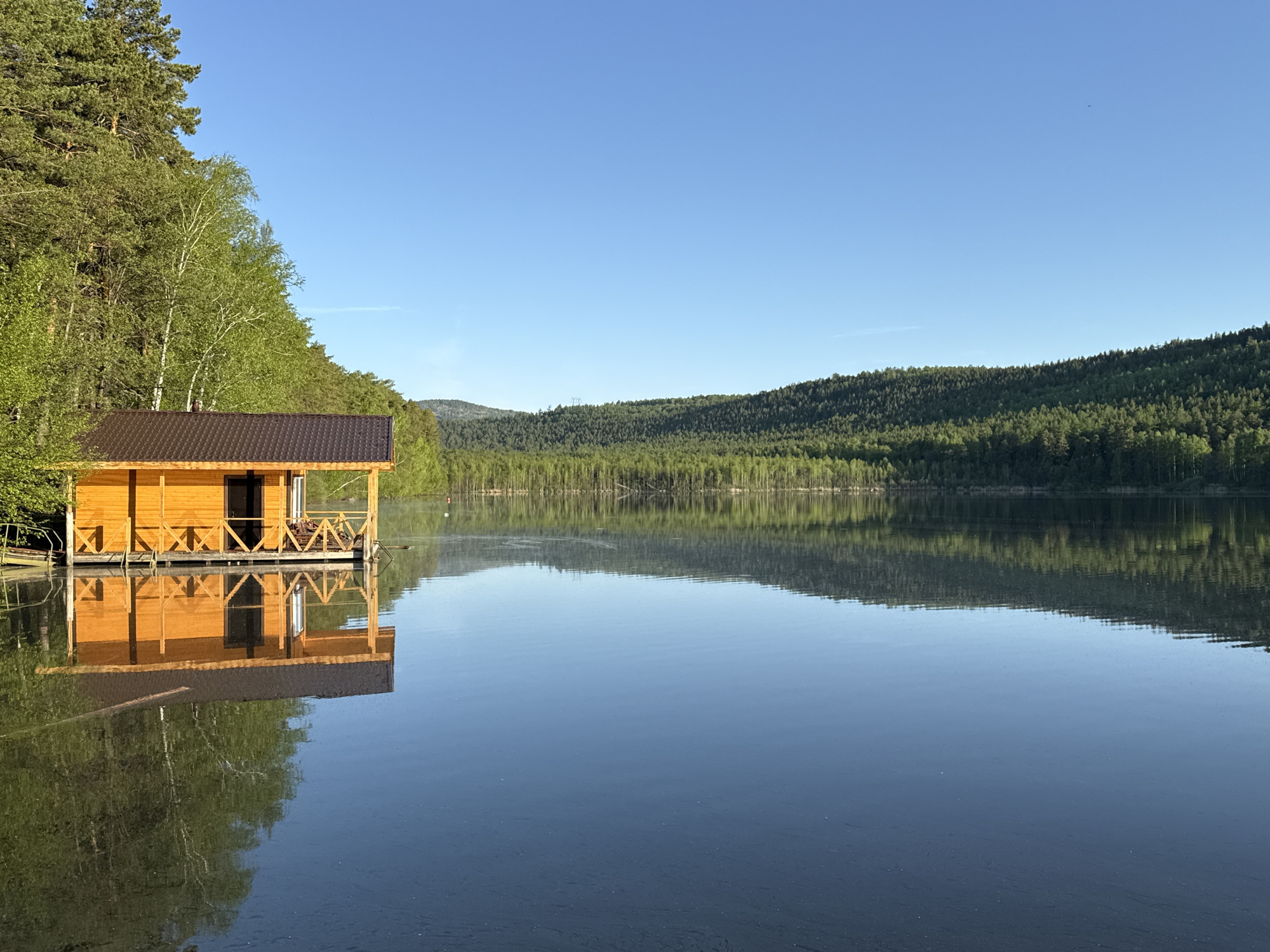 A serene landscape is captured on a clear day at Zolotoi Plyazh, Russia. A wooden cabin rests on the still water, its reflection mirrored below. Lush green trees line the shore, extending into the distance towards gentle hills. The sky is a bright, cloudless blue.