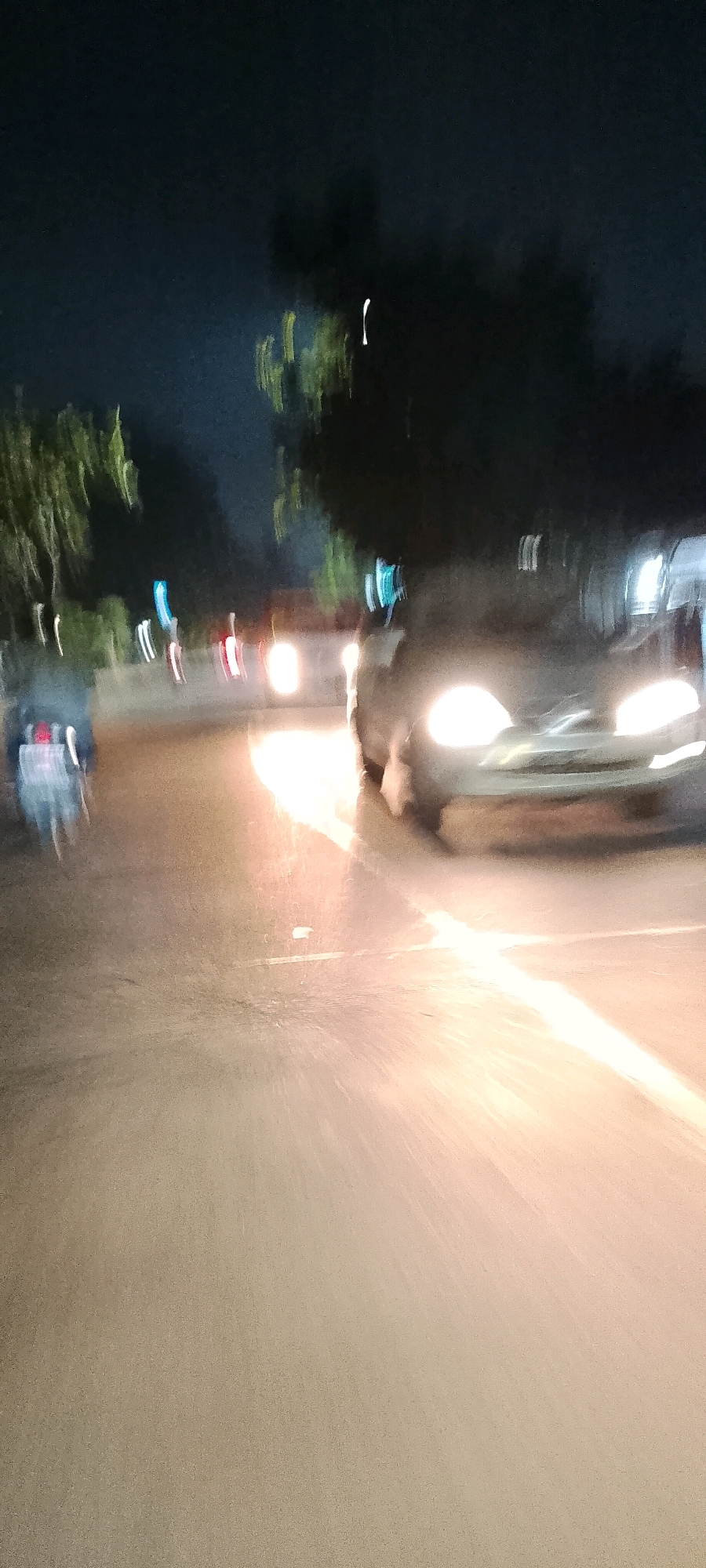 The image depicts a blurry nighttime scene on a street in Bekasi, Indonesia. The focus is on a car with its headlights on, illuminating the wet road ahead. Streaks of light from passing vehicles suggest motion blur, indicating the photograph was taken while moving or with a slow shutter speed. Trees and foliage are visible on the sides of the road, silhouetted against the dark sky. Some indistinct figures or objects are present in the background, but their details are obscured by the low light and motion blur. The overall impression is one of a busy urban street at night, characterized by artificial lighting and movement.
