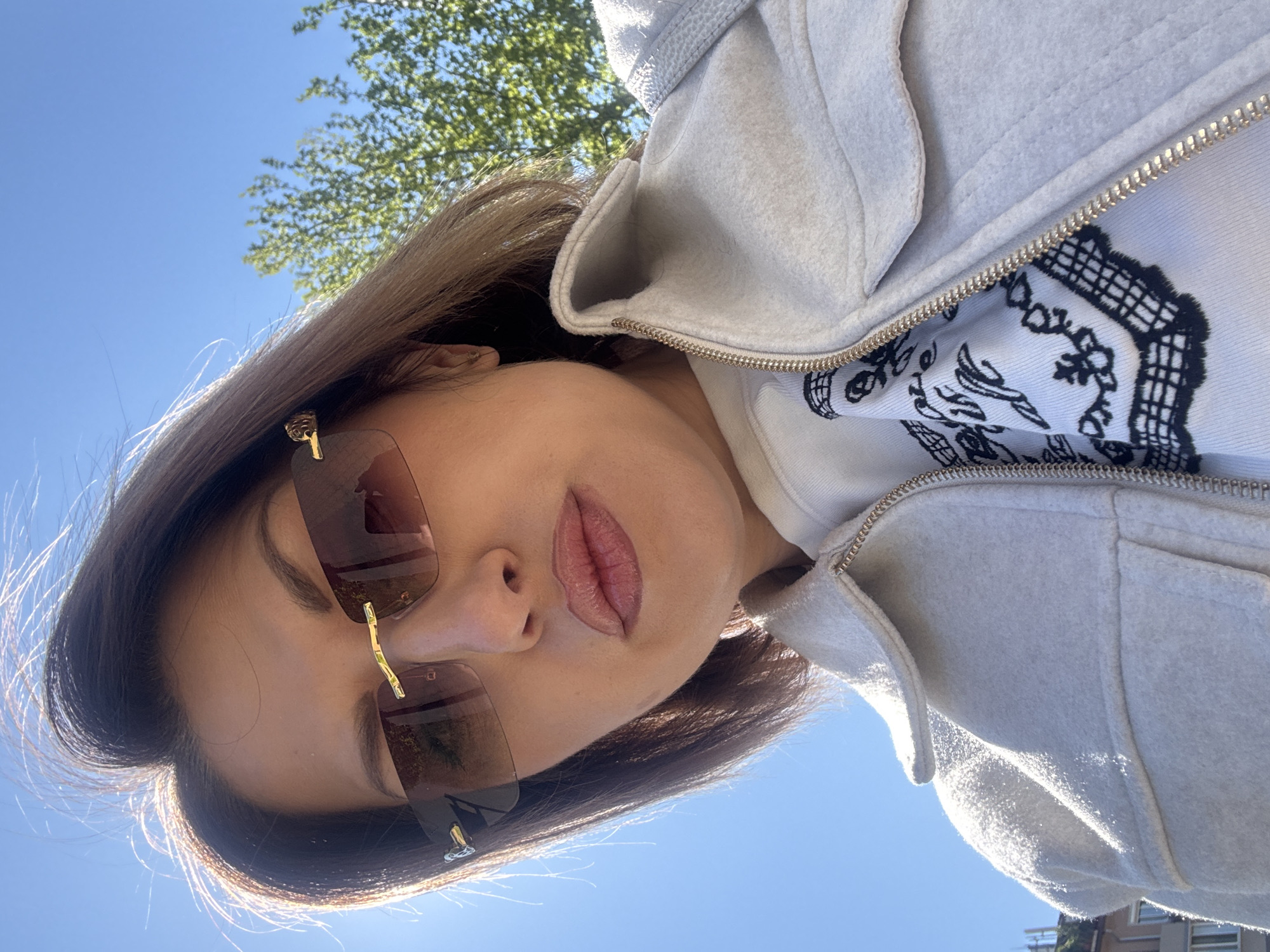 A young woman with brown hair and sunglasses is looking upwards at the camera. She is wearing a white hooded sweatshirt and a light gray zip-up jacket. The background is a clear blue sky and the green leaves of a tree. The location is not discernable as Sofia, Bulgaria from this image.