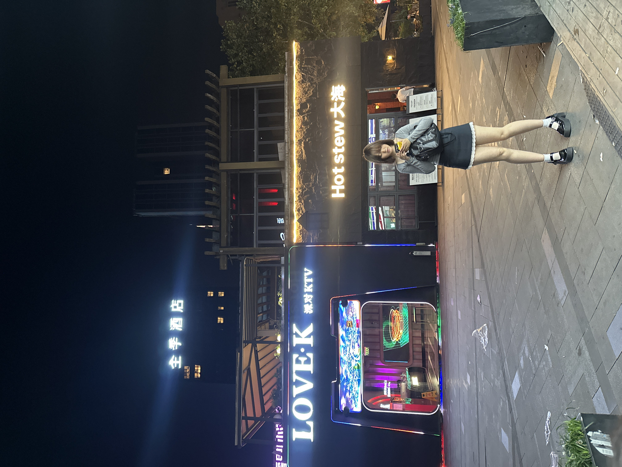 This night-time photograph captures a young woman standing on a paved pedestrian street in an urban setting, likely in Chongqing High-tech Zone, China. The image is oriented with the ground on the right and the sky on the left, suggesting it was taken with the camera rotated.
The woman, centrally positioned, wears a grey long-sleeved top, a black mini skirt with a white lace hem, white ankle socks, and black platform shoes. She carries a dark backpack and holds a drink in one hand while seemingly interacting with a phone in the other. Her gaze is directed slightly away from the camera.
The street is illuminated by various neon signs and building lights, set against a dark night sky. To the woman's left, a vibrant KTV booth with colorful LED outlines prominently displays the text "LOVE·K 迷你KTV" (Mini KTV), with glowing screens visible inside. Behind her, a building facade features a bright neon sign reading "Hot stew 大烩" (Hot Stew Big Hot Pot). Further in the background, other multi-story buildings rise, their windows showing faint interior lights. To the far left, another neon sign with Chinese characters, "扭国手艺", is visible against a dark background. The ground is paved with various shades of grey and black tiles. The overall scene depicts a lively entertainment district at night.