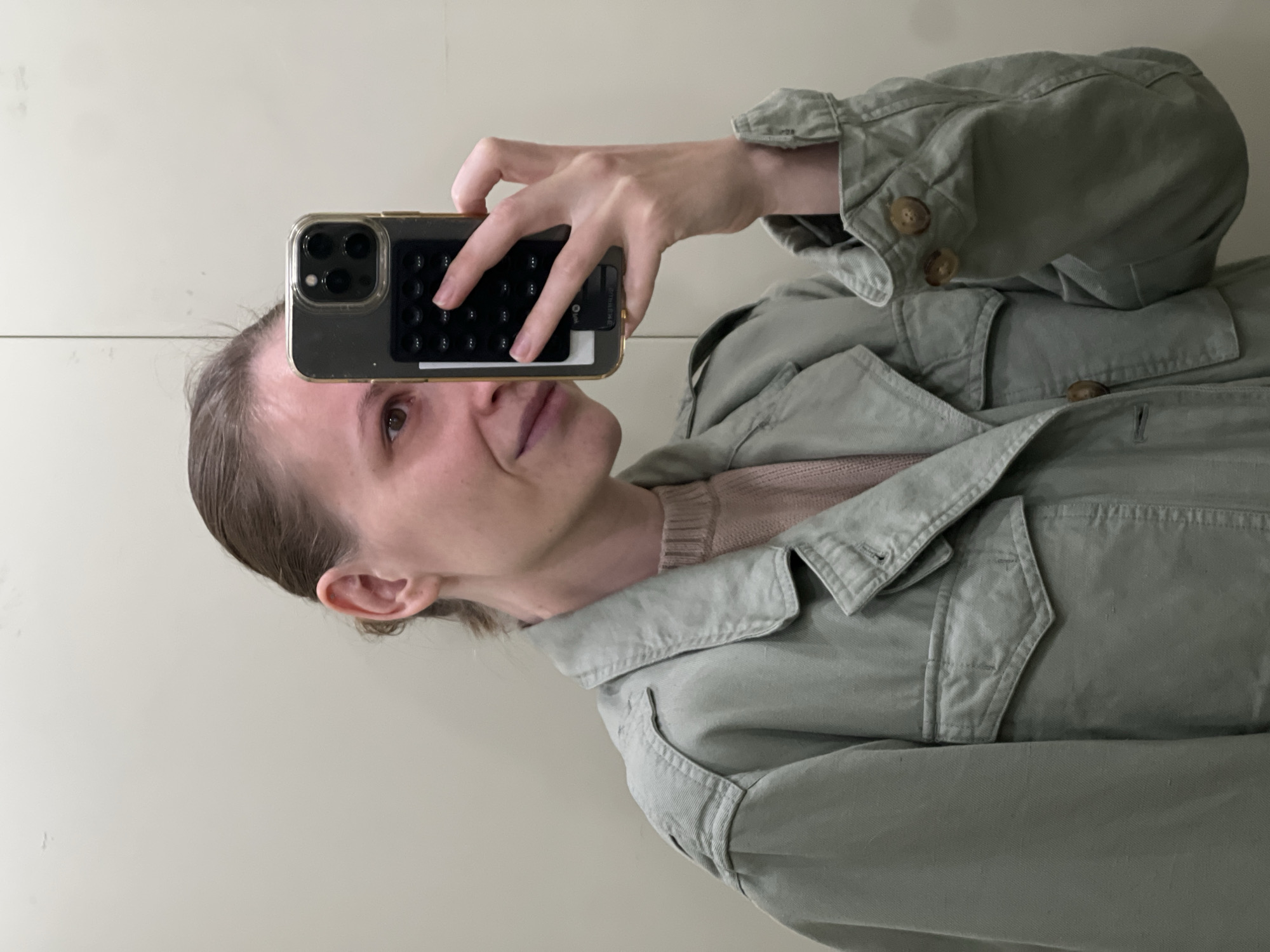 A person is taking a selfie. The person is holding a smartphone with a black keypad accessory on the back in their left hand. The person is wearing a light green jacket over a pink sweater. The background is a plain, light-colored wall. The location in Khimki, Russia is unknown.