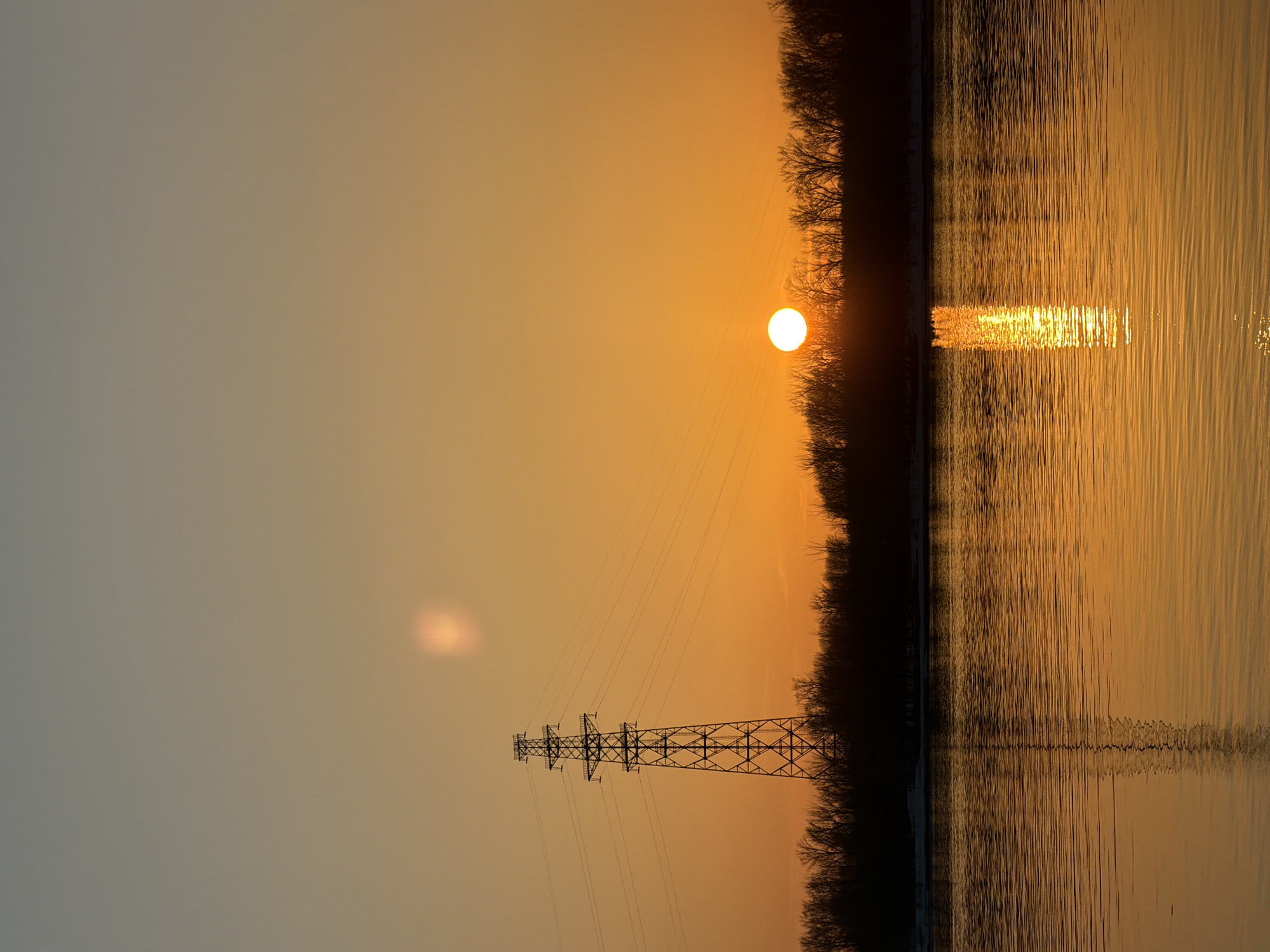 The image depicts a vertical landscape scene at sunset or sunrise, likely near the city of Chaikovsky, Russia. The scene is dominated by the golden light of the sun, which is visible in the sky and reflected in the water. A power line tower is visible, silhouetted against the warm-toned sky. A line of trees separates the sky from the water. The surface of the water is rippled, reflecting the golden light.