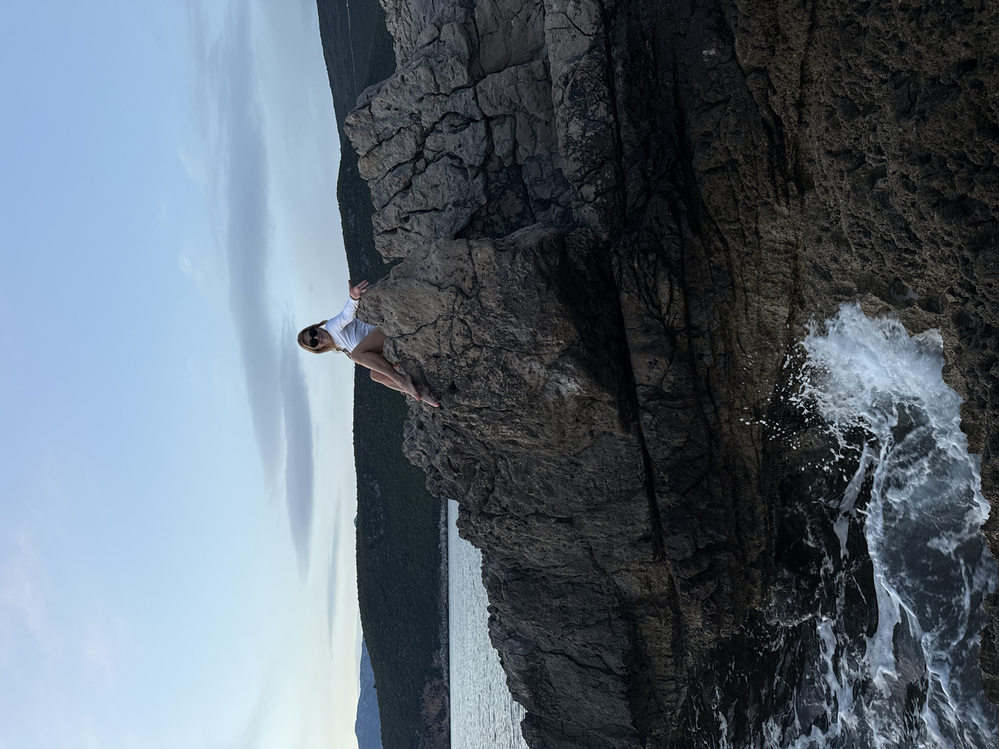 A person is sitting on a rocky promontory overlooking the sea. The scene appears to be near the coast. The location is Luštica, Montenegro. The sky is overcast and there are trees along the coast. Waves are crashing on the rocks. The person is wearing sunglasses and a white top.