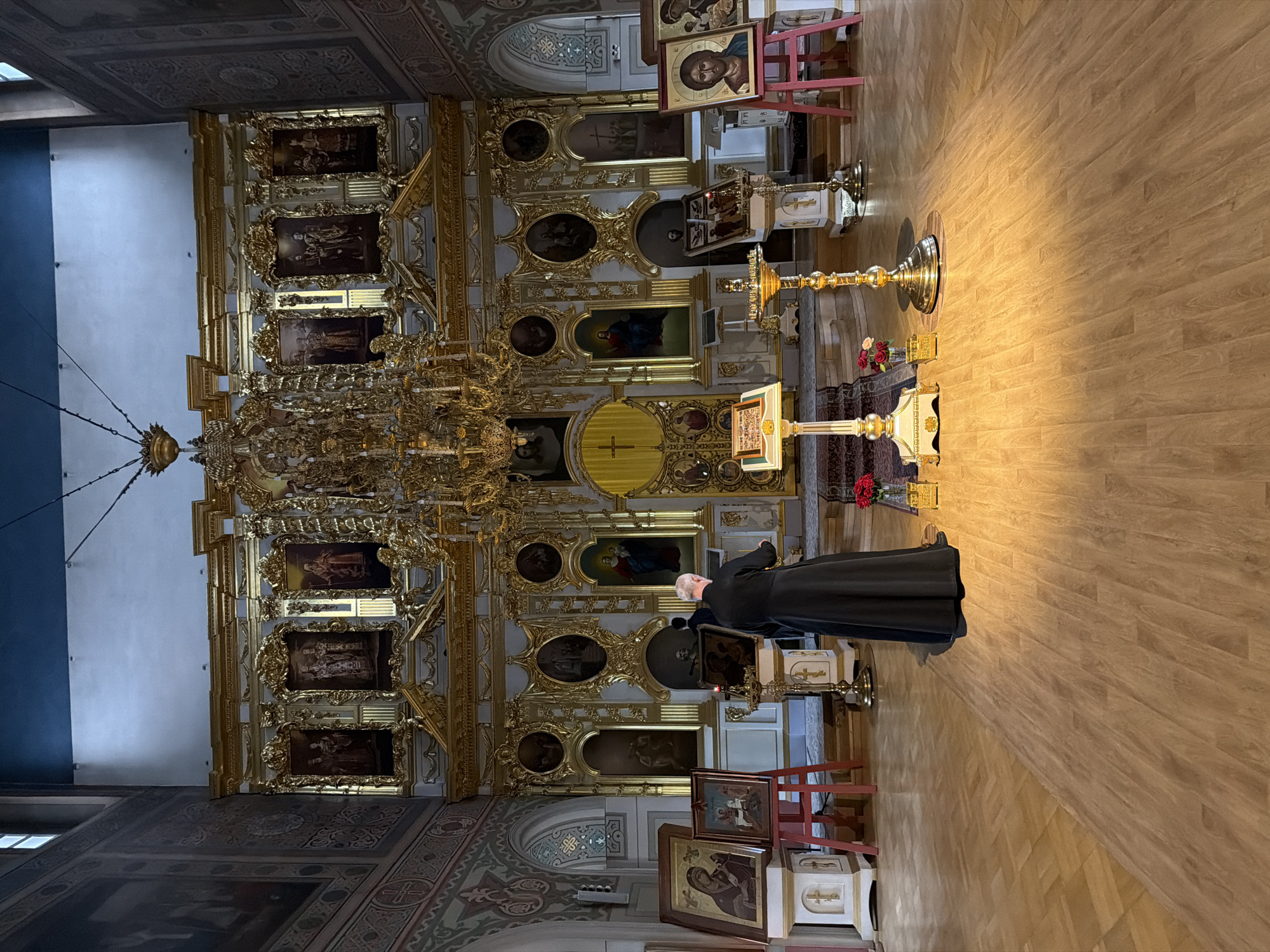 A religious figure wearing a black robe stands in the center of a church in Saint Petersburg, Russia, facing a golden iconostasis. The church interior is ornate, with gilded carvings and religious paintings adorning the walls and iconostasis. The floor is made of polished wooden parquet. Several religious icons are displayed on stands to the left and right of the central figure. The lighting is warm, casting shadows and highlighting the golden elements of the church.