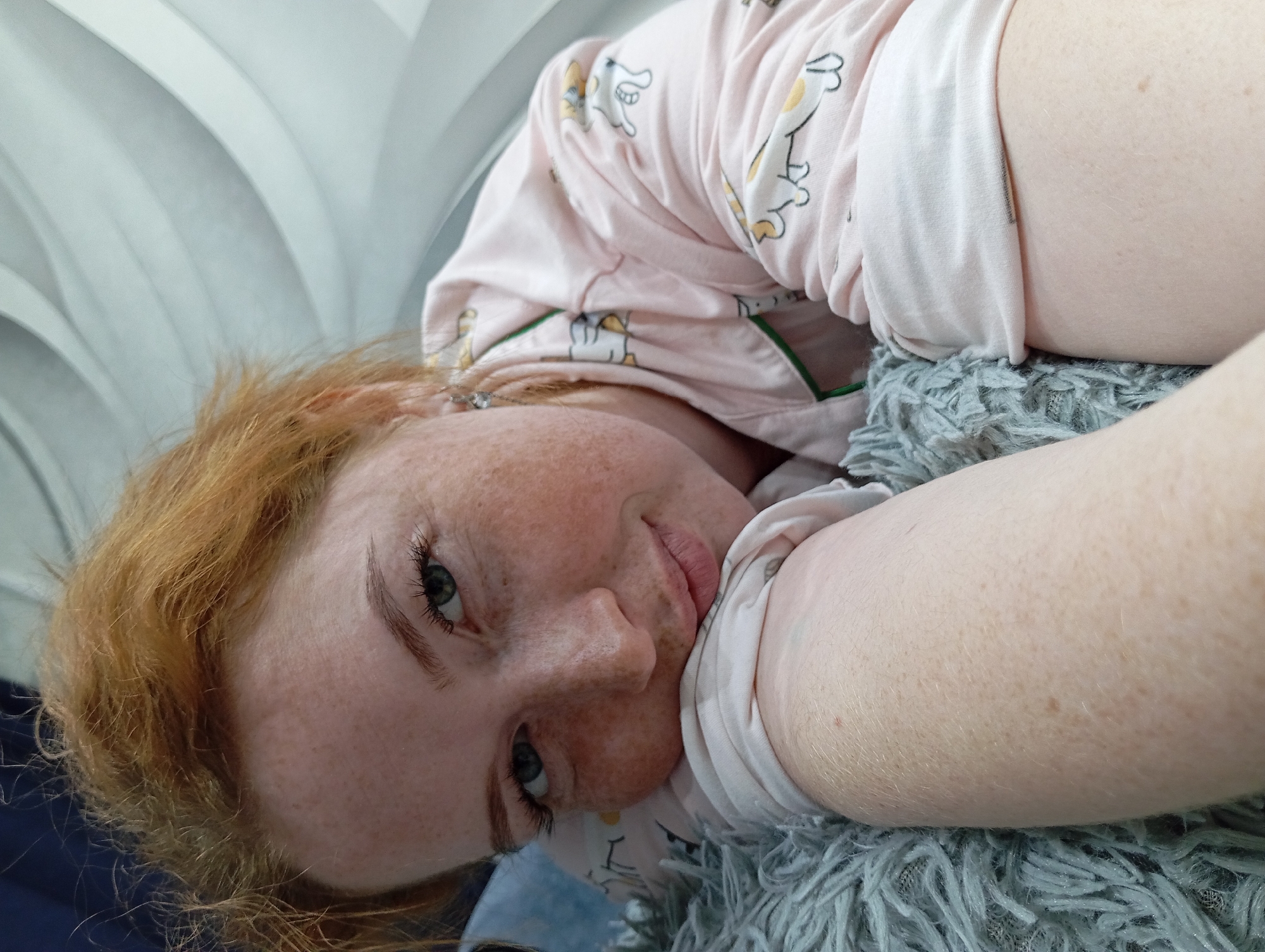 A close-up shot depicts a woman with freckled skin and reddish-blonde hair lying on a gray, textured surface that appears to be a blanket or pillow. She is wearing a light pink pajama top with a cartoon animal print. The background is partially visible and features a wavy, textured wall, possibly wallpaper or a decorative panel. The image has a slight tilt, giving a casual, relaxed feel. There is no information to suggest that the picture was taken in Yurgamysh, Russia.