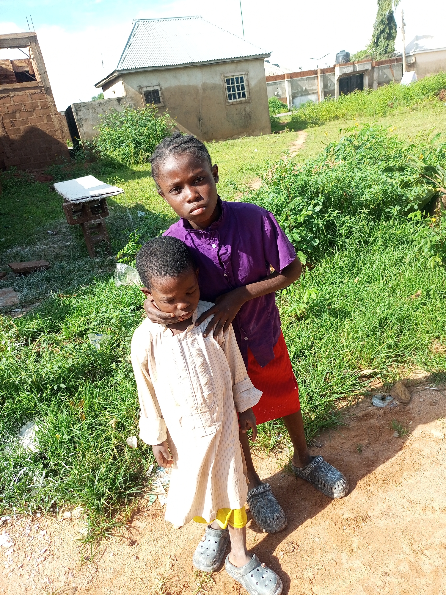In Gombe, Nigeria, two young children stand on a dirt path surrounded by green grass and scattered debris. The taller child, with dark braided hair and wearing a purple button-up shirt and a red skirt, has their arm around the shoulders of the shorter child. The shorter child, wearing a light-colored, striped tunic with yellow trim at the bottom and dark clogs, looks down with their eyes closed. In the background, unfinished and completed buildings are visible, along with a metal roof and some trees. The sky above is partly cloudy.
