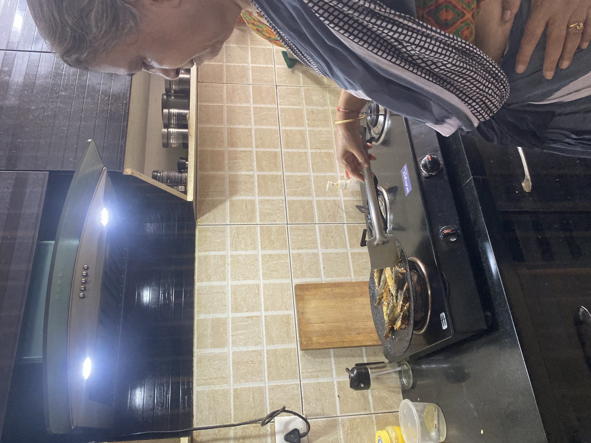 The image depicts an elderly woman cooking in a kitchen. She is using a spatula to cook small fish in a pan on a black stovetop. A range hood is visible above the stove. The kitchen has beige tiled walls and a wooden cutting board is nearby. There are jars on a shelf above the stovetop. The location is likely Chennai, India.