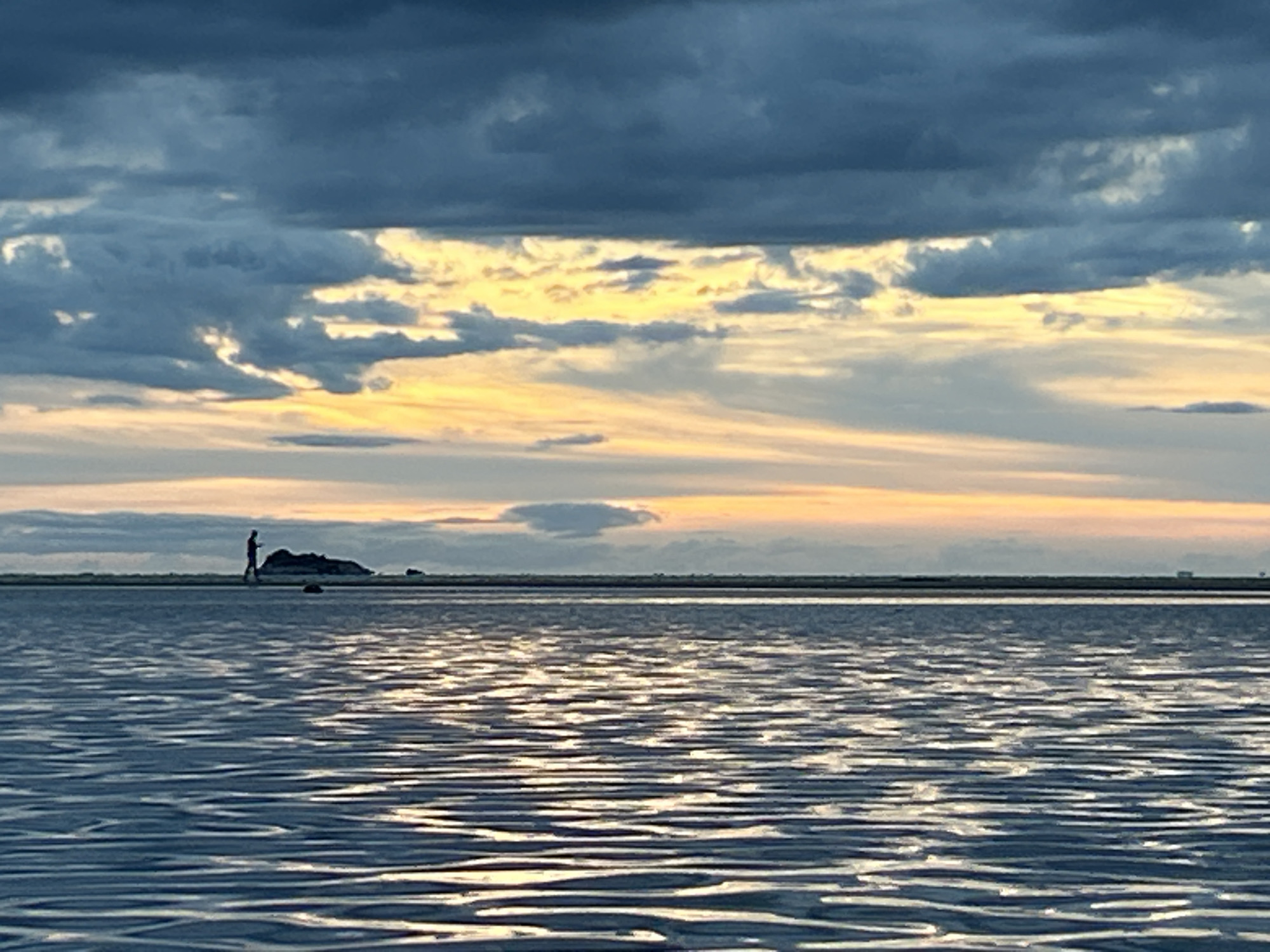 The image depicts a seascape at sunset in ตำบลเกาะพะงัน, Thailand. The foreground is dominated by rippled water reflecting the light from the sky. In the mid-ground, a person is walking on a sandbar towards a small, rocky island. In the background, the sky is filled with dark, heavy clouds with streaks of yellow and orange light breaking through, suggesting the sun is setting or has just set. The horizon line is clear, with some indistinct structures visible in the far distance. The overall mood is serene and calm.