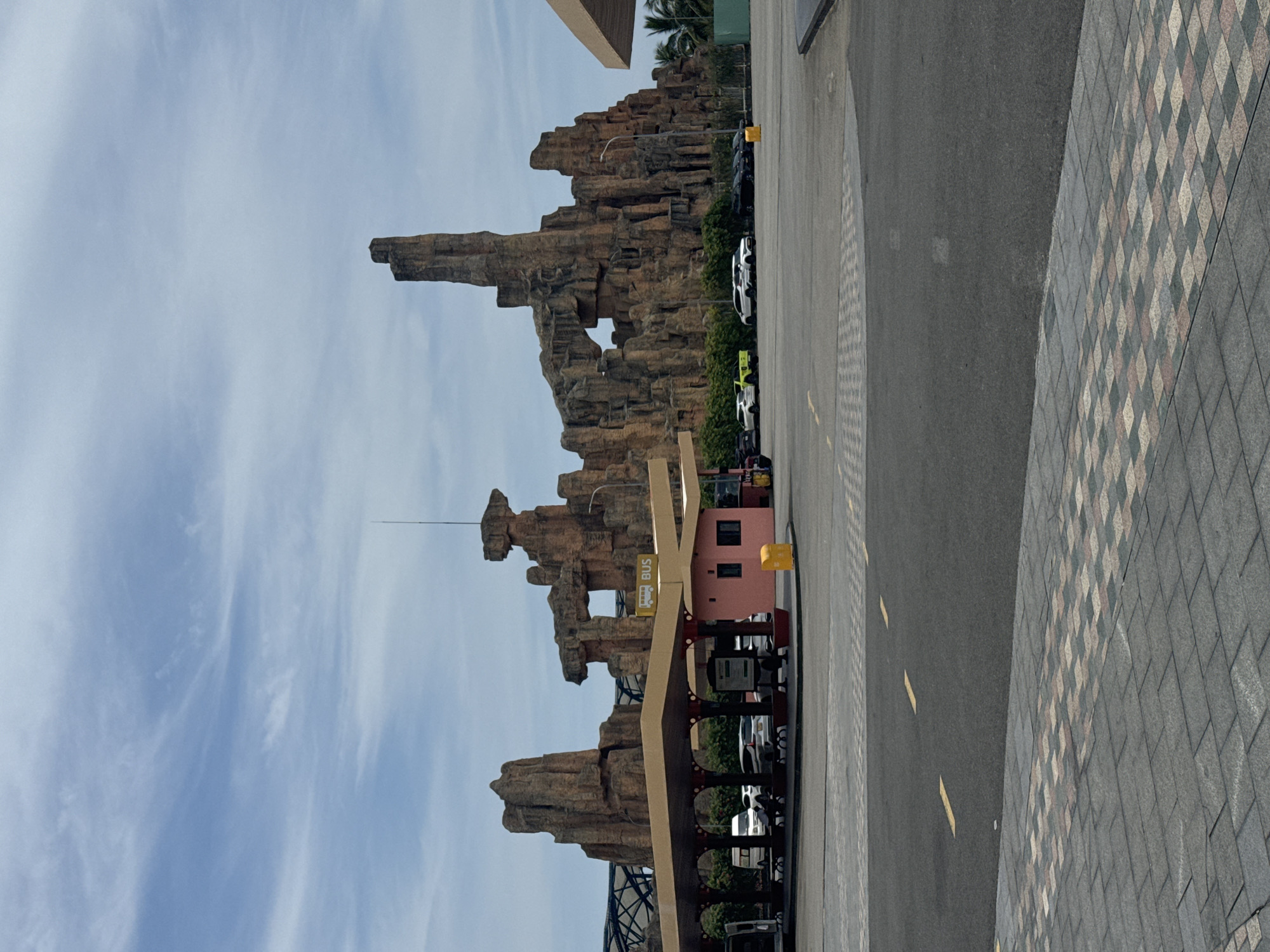 The image shows a view of the Sun World Ba Na Hills theme park in Đà Nẵng, Vietnam. The focal point is a prominent rock formation designed to resemble a mountain landscape. In the foreground is a bus stop shelter with a "BUS" sign. Various vehicles, including cars and buses, are parked nearby. The background features a partly cloudy sky. The ground surface transitions from a paved road to patterned paving stones.