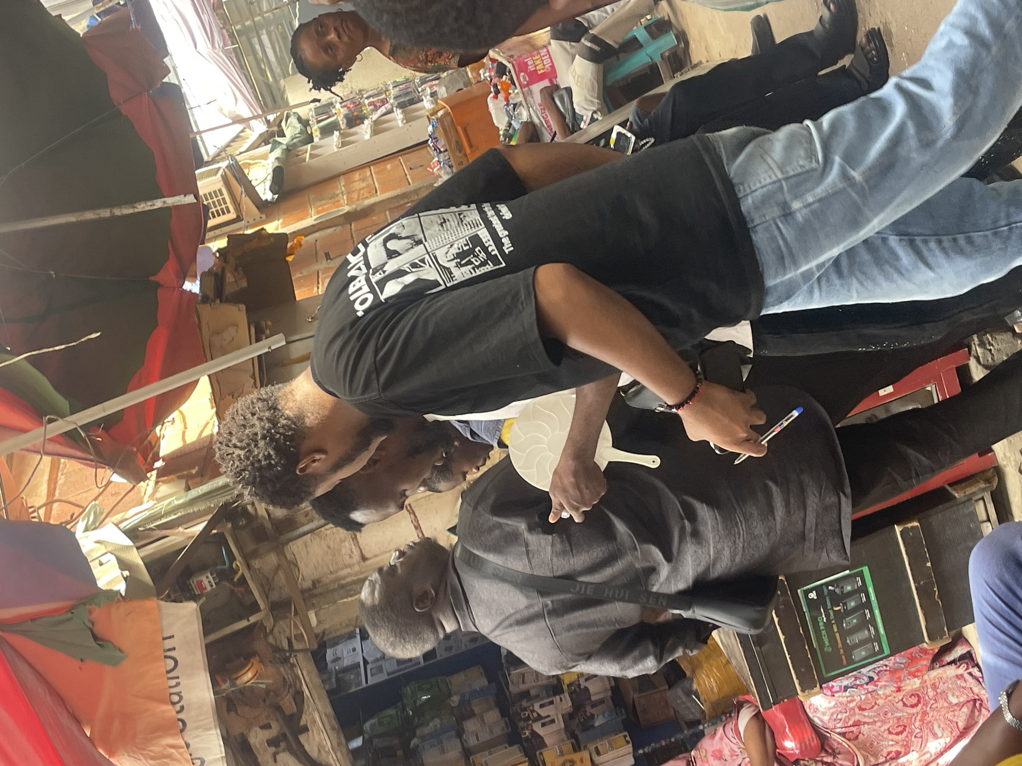 The image shows an informal market in Abuja, Nigeria. Three men are centrally featured. One man, wearing a black t-shirt and blue jeans, is standing over another man who is wearing a dark grey shirt and carrying a shoulder bag. The first man is holding a white plastic hand fan and a pen. Both men appear to be engaged in some transaction. A third person is standing in the background, watching. The market environment is crowded with stacked boxes of what appears to be retail goods. Umbrellas and makeshift shelters are visible overhead.