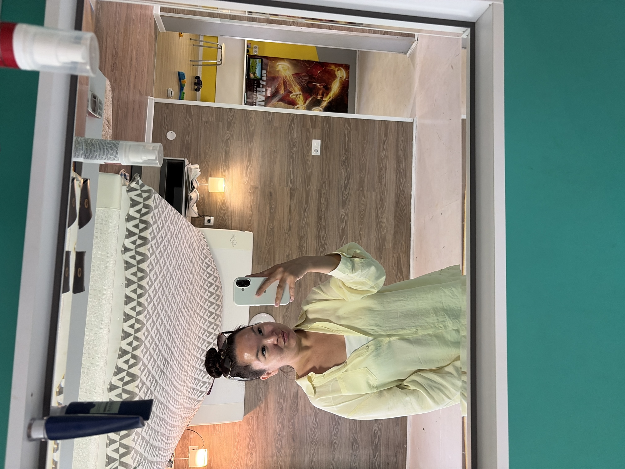 A young woman with her hair in a bun and wearing sunglasses is taking a selfie in a mirror. She is wearing a light yellow, unbuttoned, long-sleeved shirt over a white tank top. The mirror is framed by a turquoise border. The reflection shows a room with wooden walls, a patterned bedspread, and various items on shelves. To the right, a doorway leads to another room with yellow walls and a poster featuring a character from a Marvel movie. The scene appears to be an interior space, possibly a hotel room or apartment.