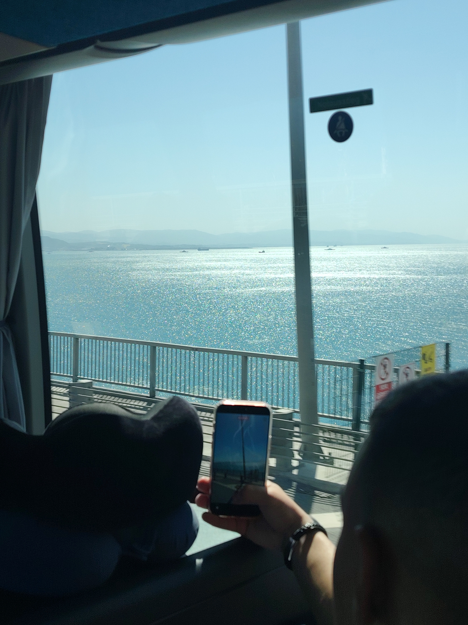 The image captures a vibrant daytime scene from inside a vehicle, likely a bus, overlooking a vast expanse of sparkling blue water, characteristic of a sea or large bay, identified as being near Dilovası, Turkey. Distant mountains line the hazy horizon under a clear, bright sky.
In the foreground, a passenger is seen holding up a smartphone to photograph the scenic view outside the window. The phone's screen displays a blue sky and what appears to be a distant structure or natural element, possibly a reflection of the exterior. The passenger's arm, adorned with a watch, and the back of their hand are visible. Another person's head and shoulder, wearing a dark garment, are partially seen in the lower right, facing away from the camera. A dark, U-shaped neck pillow rests on the seat beside the window.
Outside the window, a metal railing, part of a bridge or overpass, separates the road from the water. Several small boats are dotted across the glistening water in the middle distance. On the window itself, a dark rectangular sticker and a circular symbol, possibly indicating accessibility (like a person sitting or wheelchair user), are visible but indistinct. Two signs are attached to the railing outside; one is a red circular "no" symbol, and the other is a yellow rectangular sign, both unreadable.
The bright sunlight causing intense glints on the water indicates a sunny day, likely during midday or early afternoon, contributing to a serene and picturesque travel experience.