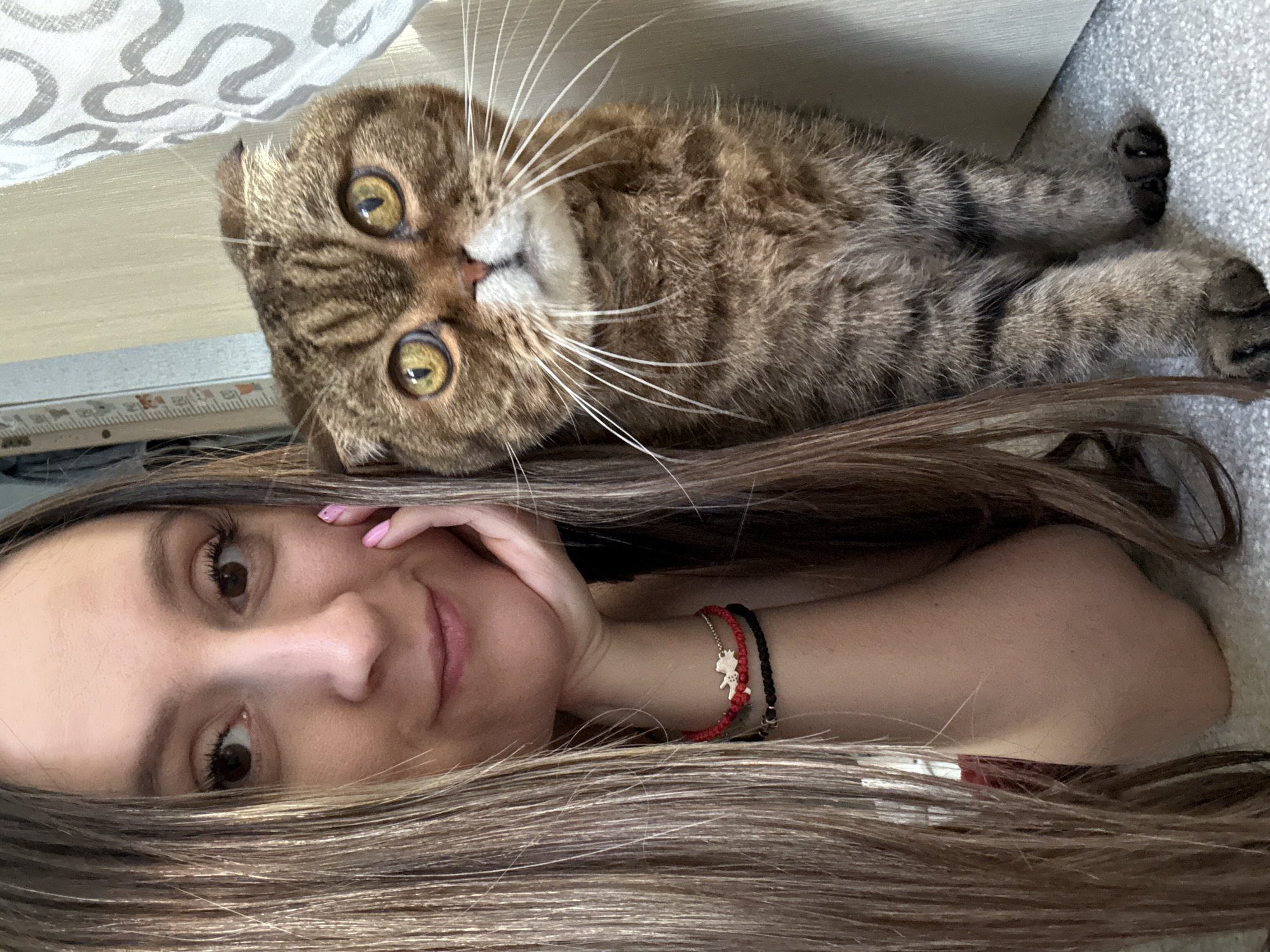The image shows a close-up selfie of a woman and a Scottish Fold cat. The woman is on the left side of the frame, with her head tilted and her hand resting on her chin. She has long, brown hair and is wearing bracelets on her left wrist. The cat is on the right side of the frame, looking directly at the camera. It is a tabby cat with distinctive folded ears and large, yellow eyes. The background appears to be indoors, with a curtain or decorative fabric visible on the right. The lighting is natural and soft. The image does not contain any elements that suggest it is located in Kroyanskoye, Russia.