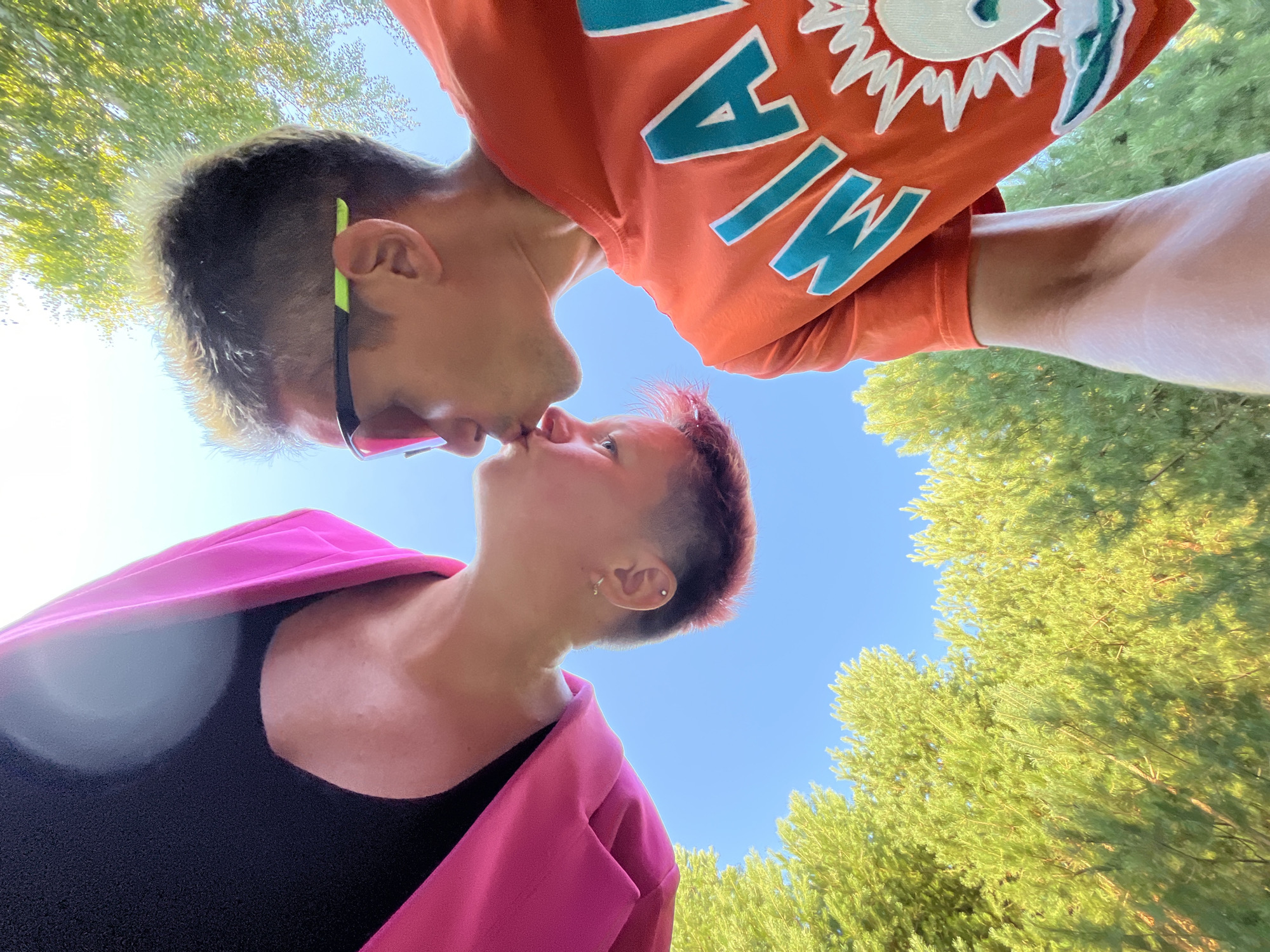 In an outdoor setting, two individuals are captured in a close-up shot, seemingly sharing a kiss. The perspective is from below, looking upwards towards the individuals and the sky. The person on the left, with short pink-dyed hair and an earring, is wearing a black top and a bright pink garment draped over their shoulder. The person on the right has styled hair with lighter tips and is wearing bright orange sports sunglasses and an orange t-shirt with a turquoise "MIA" logo and a white dolphin graphic. Both individuals are positioned against a clear blue sky, with the green foliage of trees visible in the background. The sun appears to be high in the sky, casting a bright light on the scene. There are no discernible landmarks or specific features that would indicate the city of Chaikovsky, Russia.
