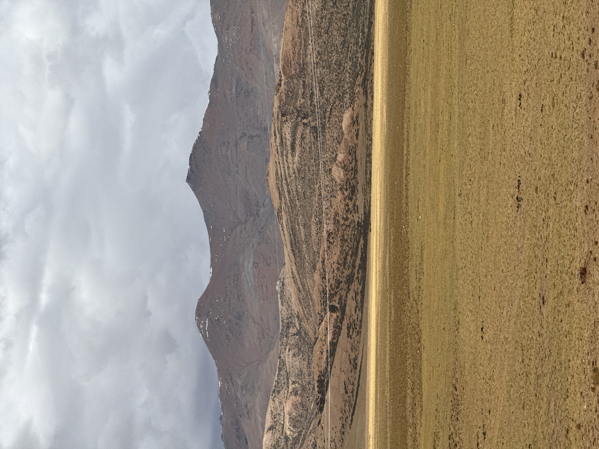 The image shows a landscape near Kela Township, China, featuring a vast expanse of golden-brown, grassy terrain. Adjacent to the field are layered mountains. The first mountain range shows brown and tan earth with vegetation. The second mountain range appears more rocky and grey. Above and behind the mountain ranges is a cloudy, overcast sky. A few power lines run along the mountain side.