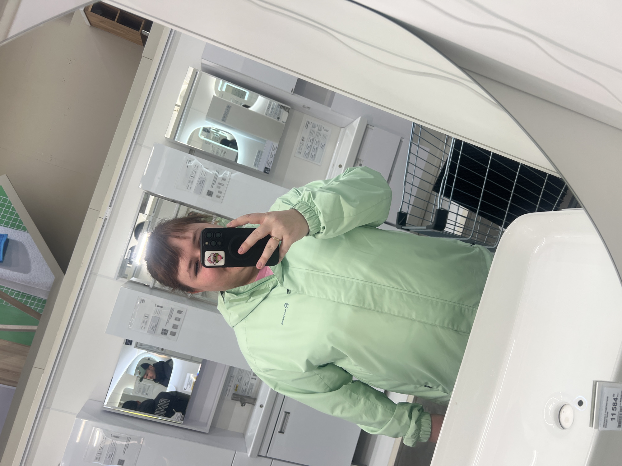 A woman wearing a light green hooded jacket is taking a mirror selfie. She is standing in what appears to be a store, possibly a home improvement store, based on the displays of bathroom fixtures and mirrors in the background. In the mirror behind the woman, there are other mirrors reflecting scenes from the store. A white sink is visible in the foreground. The label "11584" is on the sink. The location is Izhevsk, Russia.